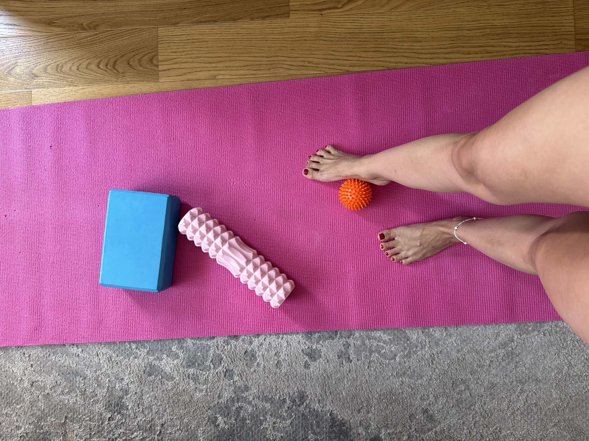 A top-down, medium shot captures a pink yoga mat on a wooden floor, bordered by a grey carpet on the left. Situated on the mat are a blue yoga block, a textured pink foam roller, and an orange massage ball. Two human feet, adorned with red nail polish and a delicate anklet on the left foot, are positioned on the mat. The right foot is near the orange massage ball, while the left foot is closer to the bottom edge of the frame. The scene appears to be indoors, possibly a home or studio, with no specific city or location details visible.