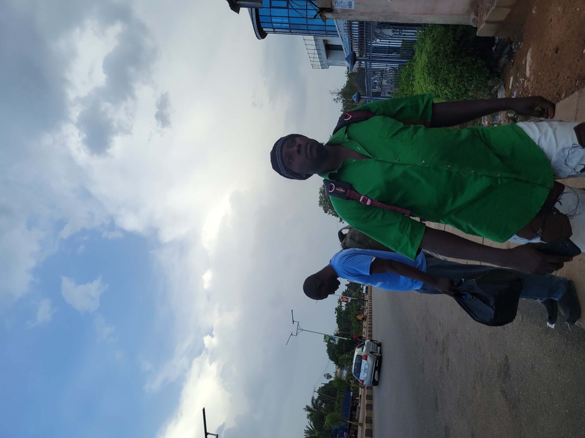 Two men are standing on the side of a paved road in Jos, Nigeria. The man in the foreground, on the right, is wearing a green button-up shirt, a black beanie, and white shorts. He has a black backpack with red straps. The man on the left is wearing a blue t-shirt and dark pants, and he is holding a black plastic bag. In the background, there are cars driving on the road, streetlights, trees, and a modern building with blue windows. The sky is cloudy.