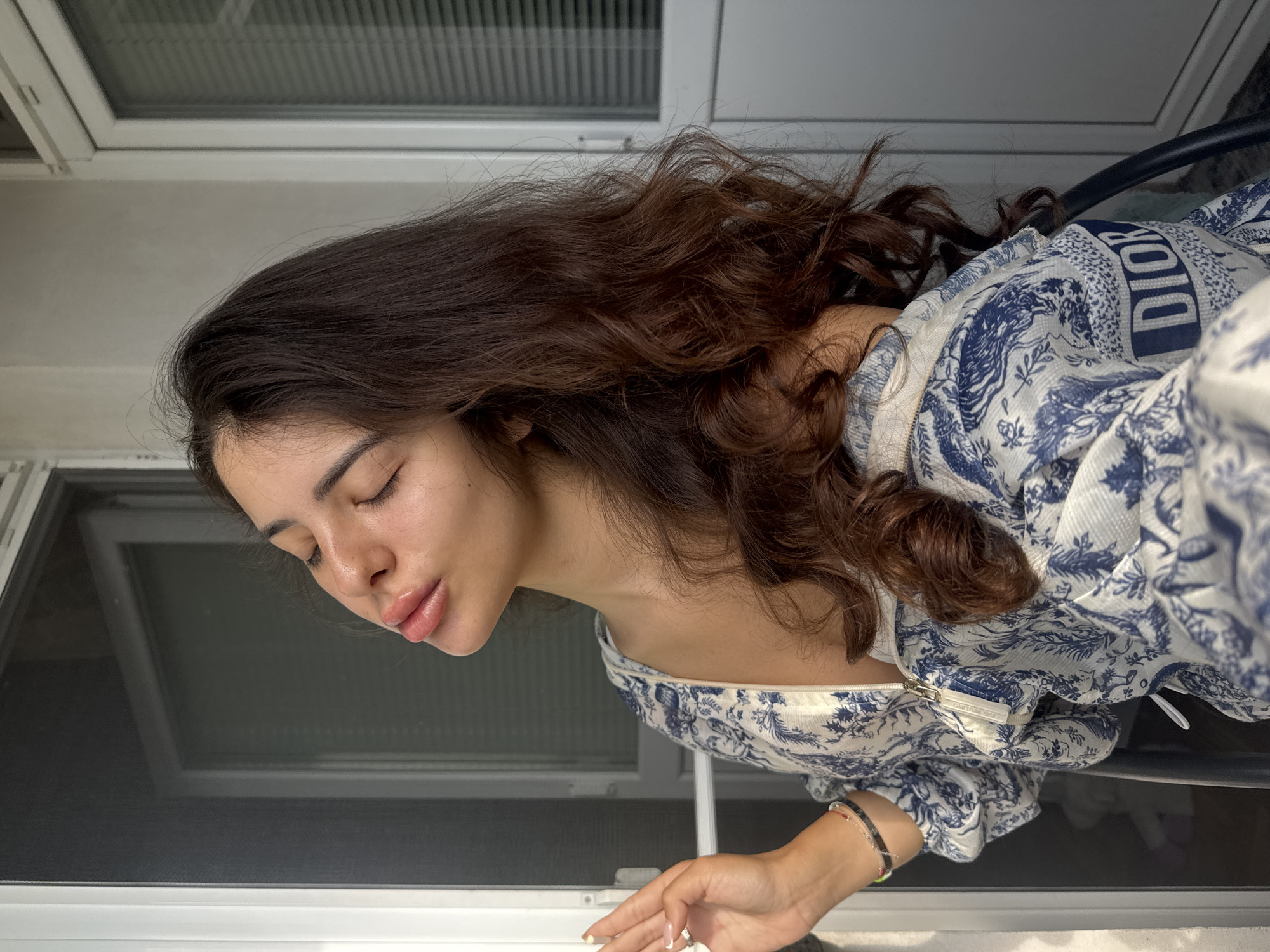 A young woman with long, dark, wavy hair is shown from the chest up, sitting outdoors. She is wearing a white top with a blue floral pattern and a zipper. Her eyes are closed, and her lips are slightly parted, as if she is puckering. Her head is tilted back, and she is looking upwards. Her left hand is raised, with her fingers slightly curled, near a window screen. The background consists of a white wall and a window with blinds. The scene appears to be on a balcony or patio, likely in a residential area. No specific landmarks of Plovdiv, Bulgaria are visible.