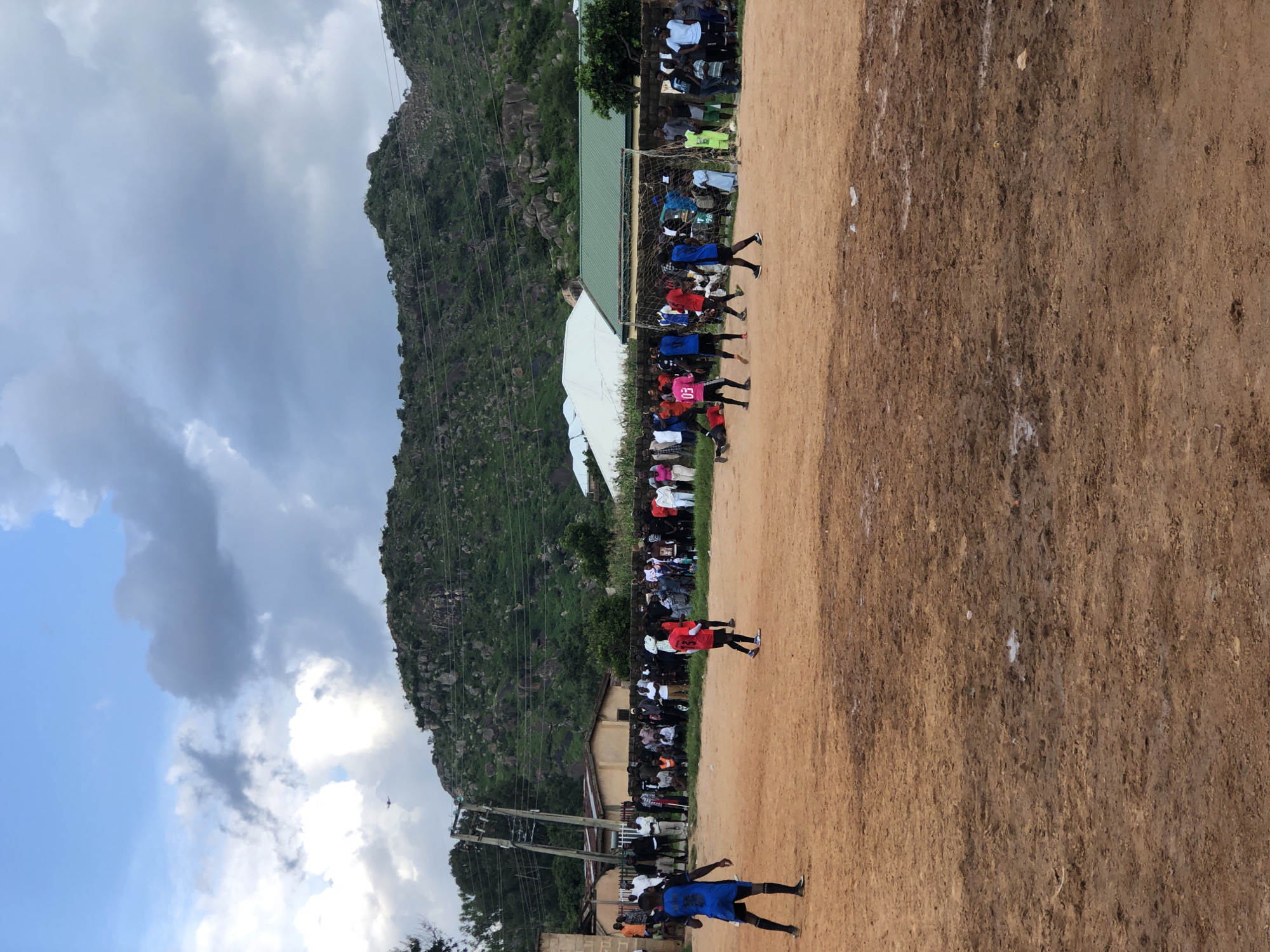 In Jos, Nigeria, a soccer match is underway on a muddy, dirt field. A group of young men, some wearing blue and red jerseys, are playing a game. Spectators, dressed in various casual attire, are gathered around the perimeter of the field, watching the game. In the background, a line of buildings, including structures with white and green roofs, are visible. Behind the buildings, a rocky, green hillside rises sharply, with a cloudy sky overhead. The scene depicts a local sporting event in a community setting.