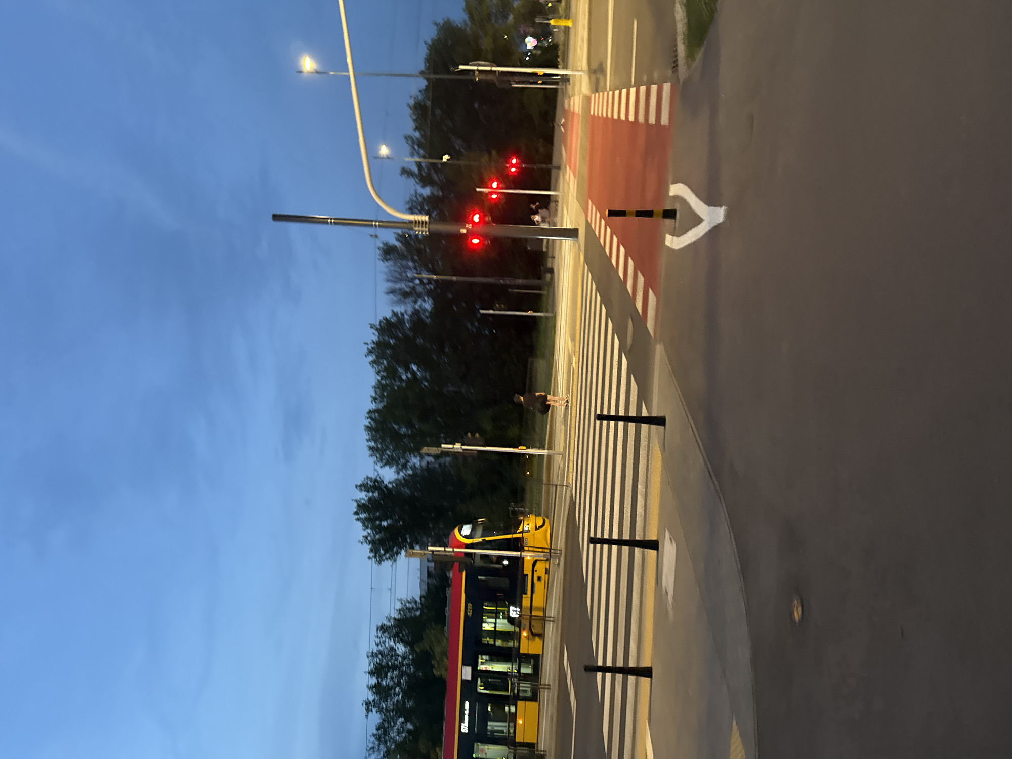 A low-angle, full shot captures a city street scene at dusk. The sky is a deep, cloudy blue, transitioning into darkness. In the foreground, asphalt paving is visible with white directional arrows painted on it, pointing away from the viewer. To the left, a black and yellow striped bollard stands near a yellow hatched area. Further left, a pedestrian crossing with white stripes and yellow edges spans the road. A yellow tram with a red roof is stopped at the crossing, its doors closed, and passengers visible inside. A single person is walking across the pedestrian crossing, away from the tram, wearing dark clothing and carrying a bag. Across the street, traffic lights display red signals. Trees line the background, and a lamppost with a bright light is visible on the right.