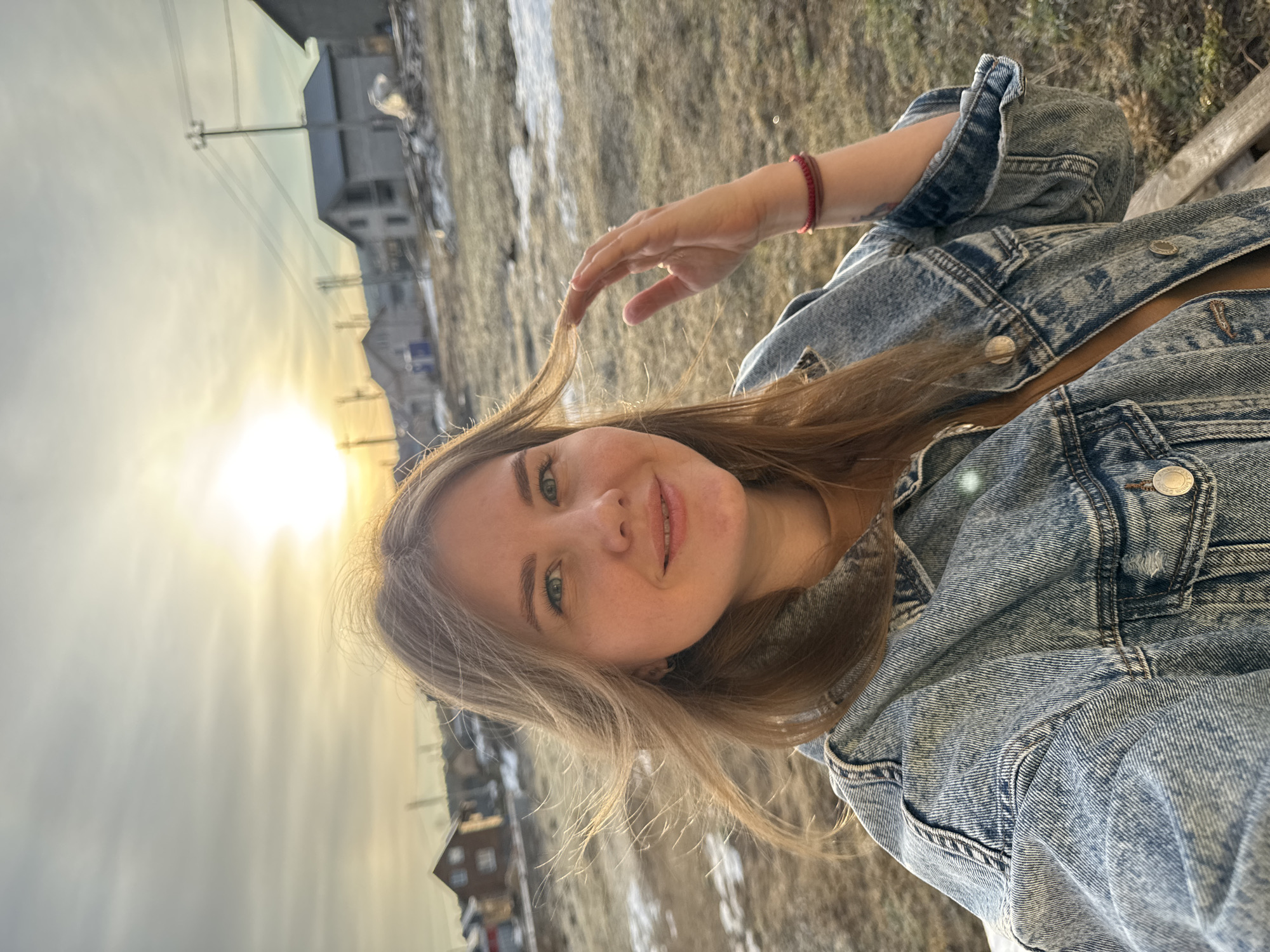 A young woman with light blonde hair is smiling in a selfie shot, wearing a denim jacket. She is sitting on a bench. The sun is shining brightly in the background, illuminating the sky and the surrounding landscape. Behind her are some houses in the city of Ферма, Russia. The ground appears to be a mix of grass and patches of snow.