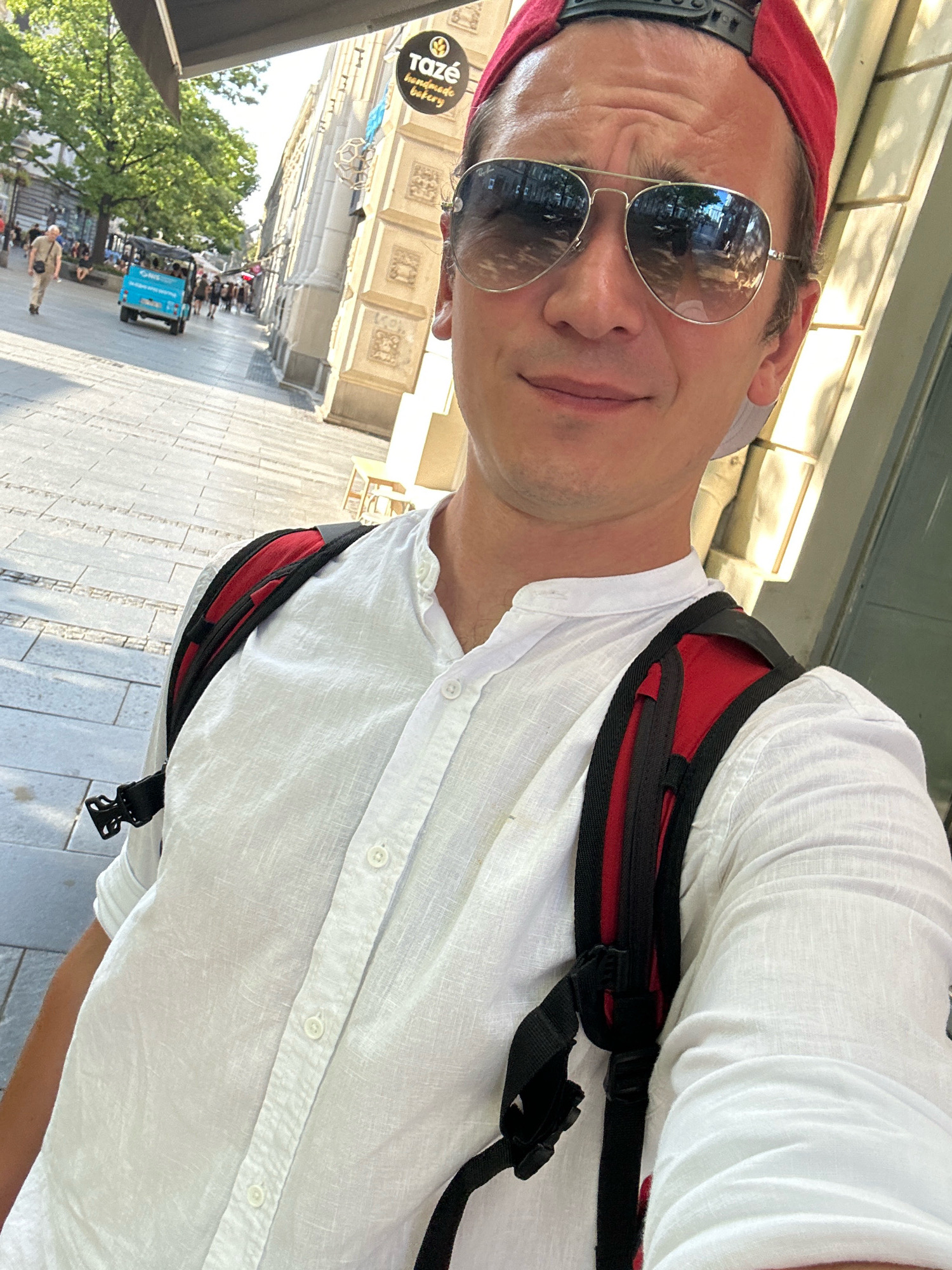 A man wearing a white linen shirt, a red baseball cap, and aviator sunglasses takes a selfie outdoors on a sunny day. He is carrying a backpack with red accents. In the background, a cobblestone street is visible, lined with buildings and trees. A small blue vehicle, possibly a tourist shuttle, is driving down the street, and several pedestrians are walking. A sign for "taze handmade bakery" is also visible on one of the buildings. The scene appears to be in a pedestrian zone or a busy street in the city of Belgrade, Serbia, likely during warmer months.