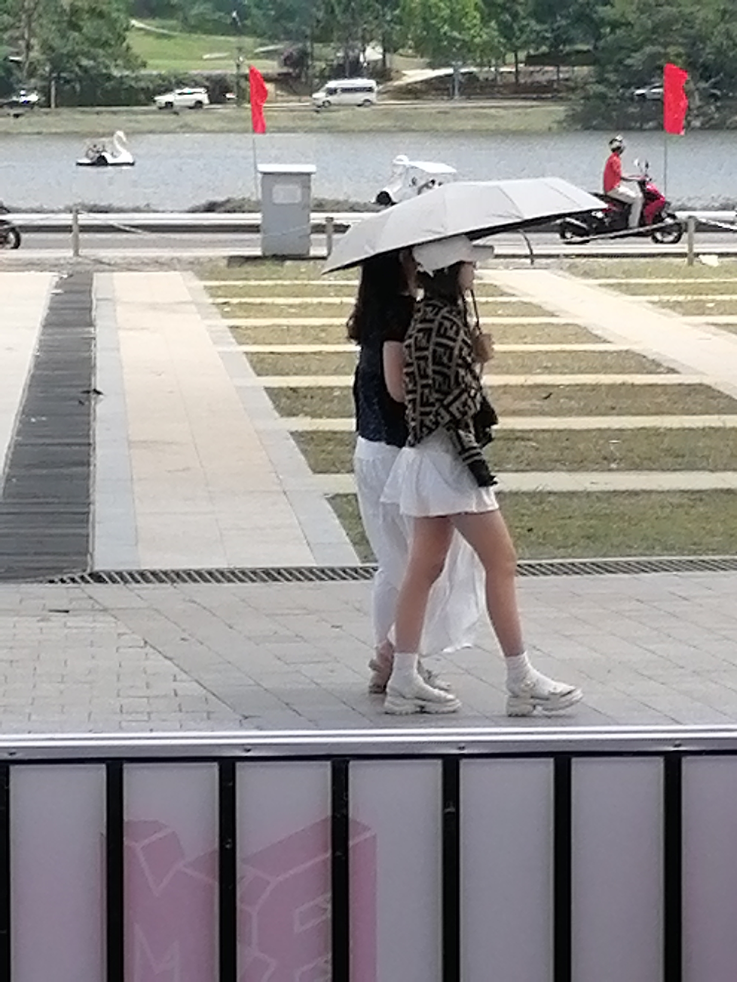The image captures a street scene in Da Lat, Vietnam, with two individuals walking side-by-side under a large umbrella. The person on the left is wearing a black top and white pants, while the person on the right is wearing a patterned jacket and a white skirt. Both are wearing white socks and chunky white sneakers. They are walking on a paved walkway with a drainage grate. In the background, there is a body of water with a swan-shaped pedal boat, a white van, a red flag, and a man riding a red motorcycle. The environment suggests a park or recreational area, possibly near a lake, given the presence of water and lush greenery. The lighting indicates it is daytime.