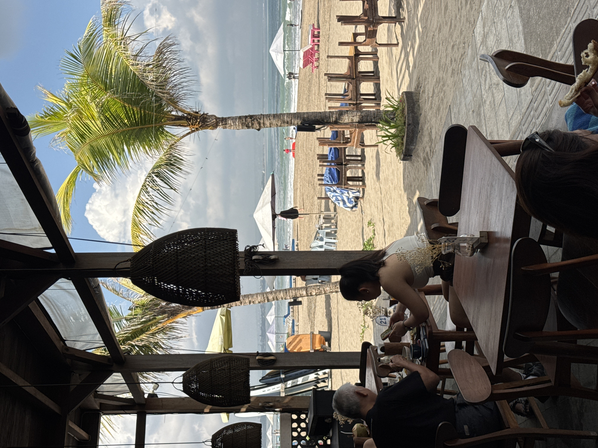 The image captures a lively scene from an open-air beachfront restaurant or cafe, offering a view of a sandy beach and the ocean, likely in Kuta, Indonesia.
In the foreground, within the shaded establishment, wooden tables and chairs are arranged. Two individuals are seated at a table: a woman with dark hair, wearing a white top with lace details, leans over the table, seemingly focused on an item in her hands. Beside her, a man with grey hair in a black t-shirt is also looking down at the table. On their table, a small vase containing dried flowers and a white circular sign with the number "21" are visible. To the far right, another person's head and shoulder are seen, holding what appears to be a piece of bread or food. The restaurant's ceiling features exposed wooden beams and several dark, woven basket-like light fixtures hanging down.
Beyond the restaurant's structure, a broad, sunlit sandy beach stretches out. A prominent palm tree stands centrally, its fronds reaching towards a bright blue sky with scattered white clouds. The beach is dotted with rows of wooden lounge chairs and numerous white and light-colored beach umbrellas. A blue and white striped towel rests on one of the lounge chairs, and an orange portable water dispenser is visible further back on the sand.
Towards the horizon, the ocean meets the shore with gentle waves. Several distant figures are scattered along the beachfront and in the water, appearing to be engaged in activities like walking, swimming, or surfing. The time of day appears to be late morning or early afternoon, with clear, sunny weather. The overall setting evokes a relaxed and leisurely tropical vacation atmosphere.