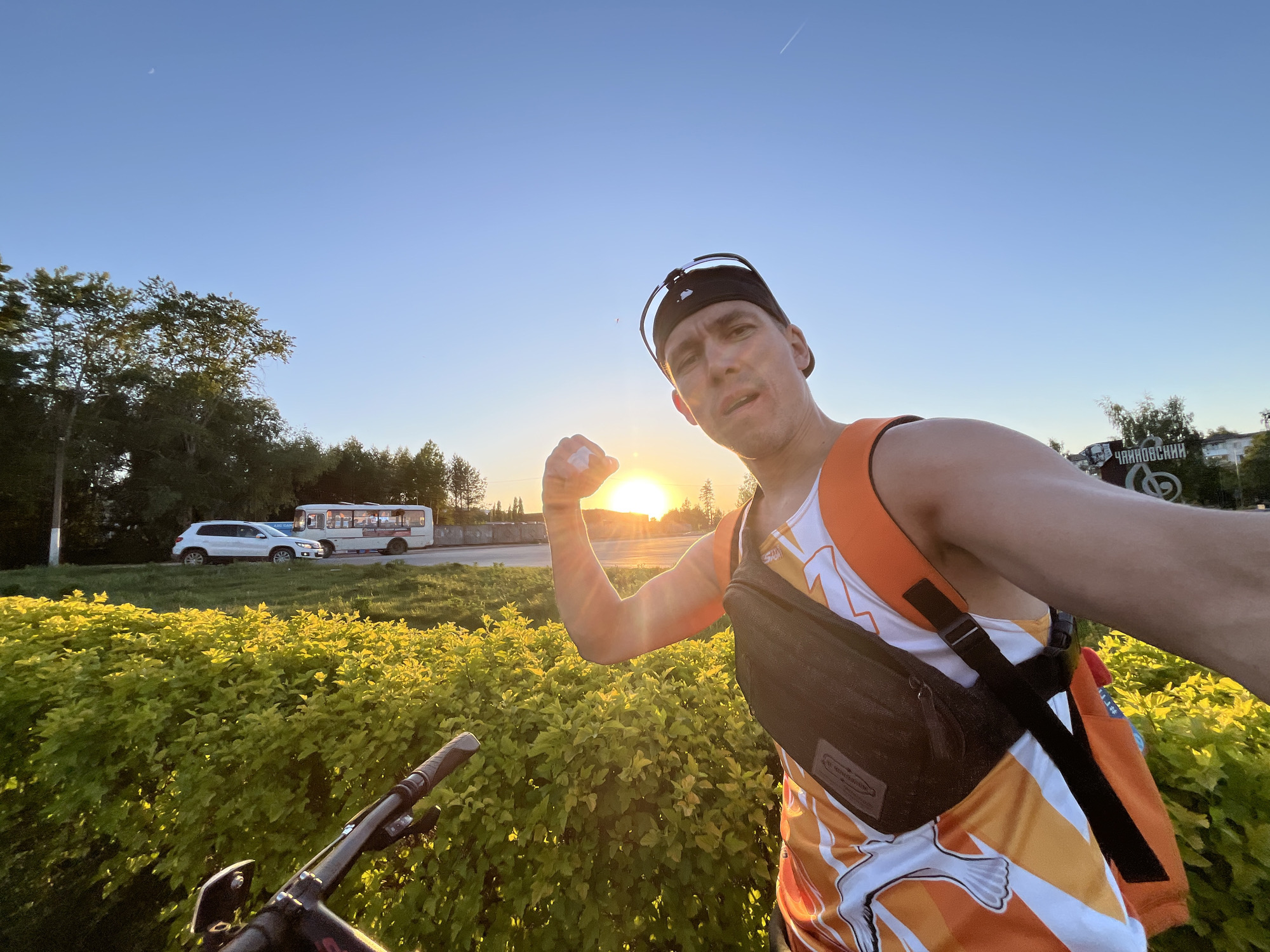 A selfie shows a man with short brown hair, wearing a black visor, an orange and white tank top, and a black fanny pack across his chest. He is flexing his right arm towards the camera, with the sun in the background. The background includes a low green shrubbery and a street with a white SUV and a white bus. The street sign in the background reads "Чайковский" (Chaikovsky in Russian). The sky is light blue. The lower part of a bicycle is visible in the lower left corner.