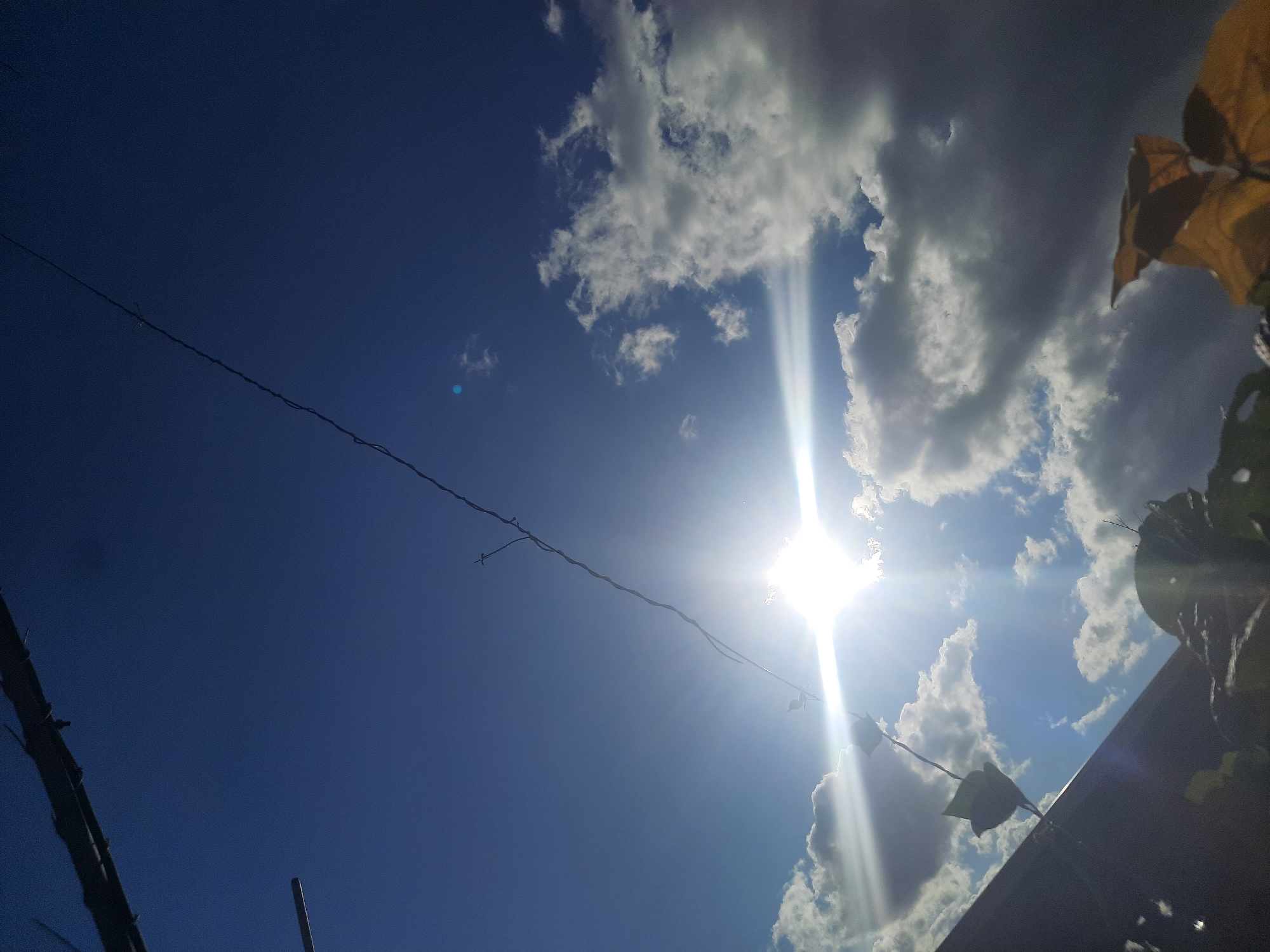 This image, taken in Shklov, Belarus, is an upward-facing shot of a bright, partly cloudy sky. The sun is intensely shining, creating lens flares that extend across the frame, particularly from the lower left quadrant. A dark, thin wire stretches diagonally from the upper left to the lower right of the image. In the foreground, obscuring the bottom portion of the frame, are dark silhouettes of foliage and what appears to be the edge of a dark roof or structure. The sky is a deep blue, punctuated by fluffy white and gray clouds, some of which are illuminated by the sun. There are no people or animals visible in the image.