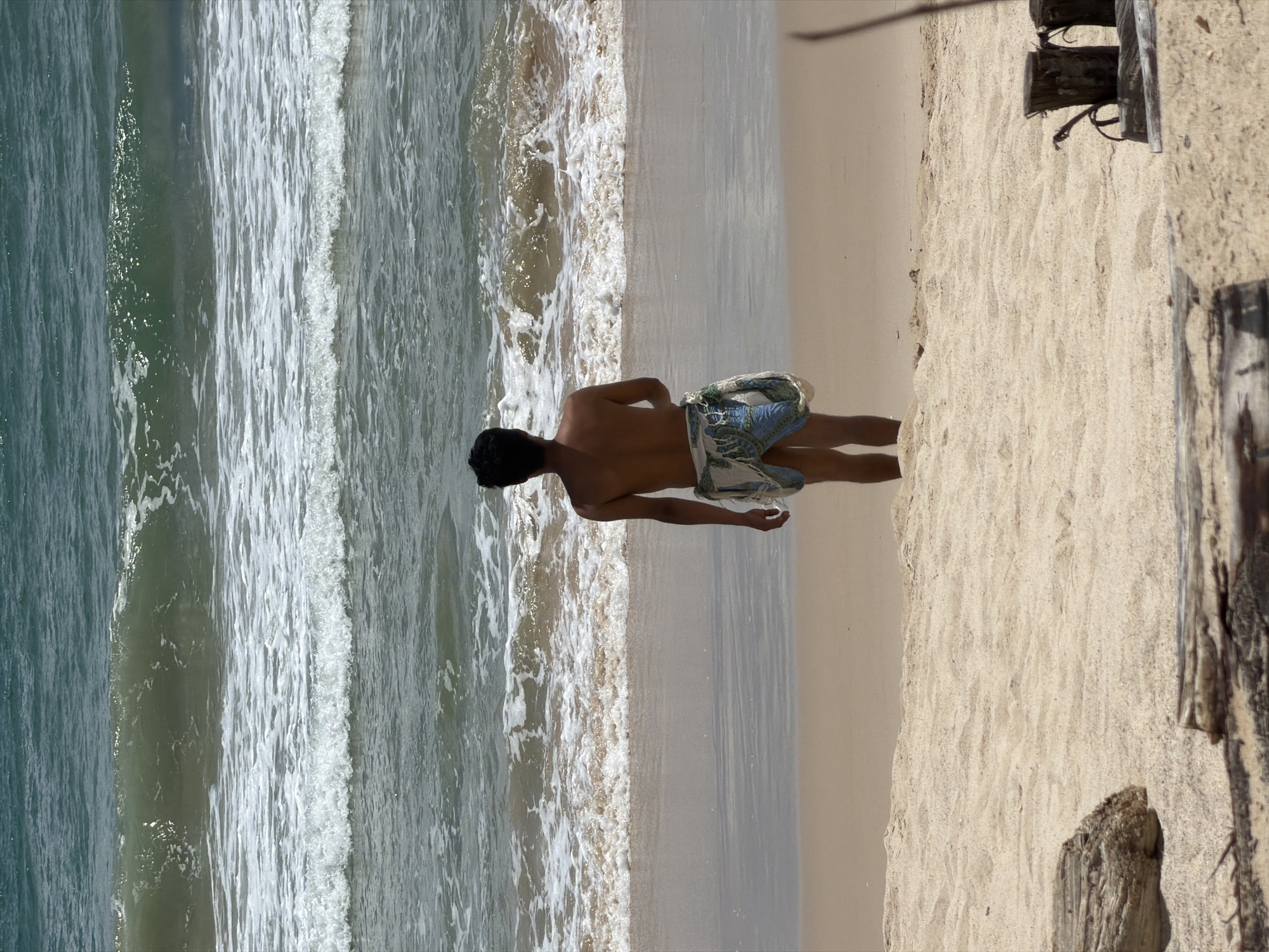 The media file depicts a young man standing on a sandy beach, with the ocean and waves to his left. He is wearing patterned swim shorts and is facing away from the camera, looking out towards the sea. The water appears to be a clear, greenish-blue, with gentle waves breaking on the shore. The sand is light brown and appears to be dry and slightly rippled. In the background, there are some weathered wooden structures, possibly part of a pier or boardwalk, suggesting a developed coastal area. The lighting suggests it is daytime, likely sunny, casting shadows on the sand and highlighting the texture of the waves. The overall scene evokes a sense of relaxation and enjoyment of a tropical beach environment. The location context of Choeng Thale, Thailand, aligns with this image of a sunny beach destination.