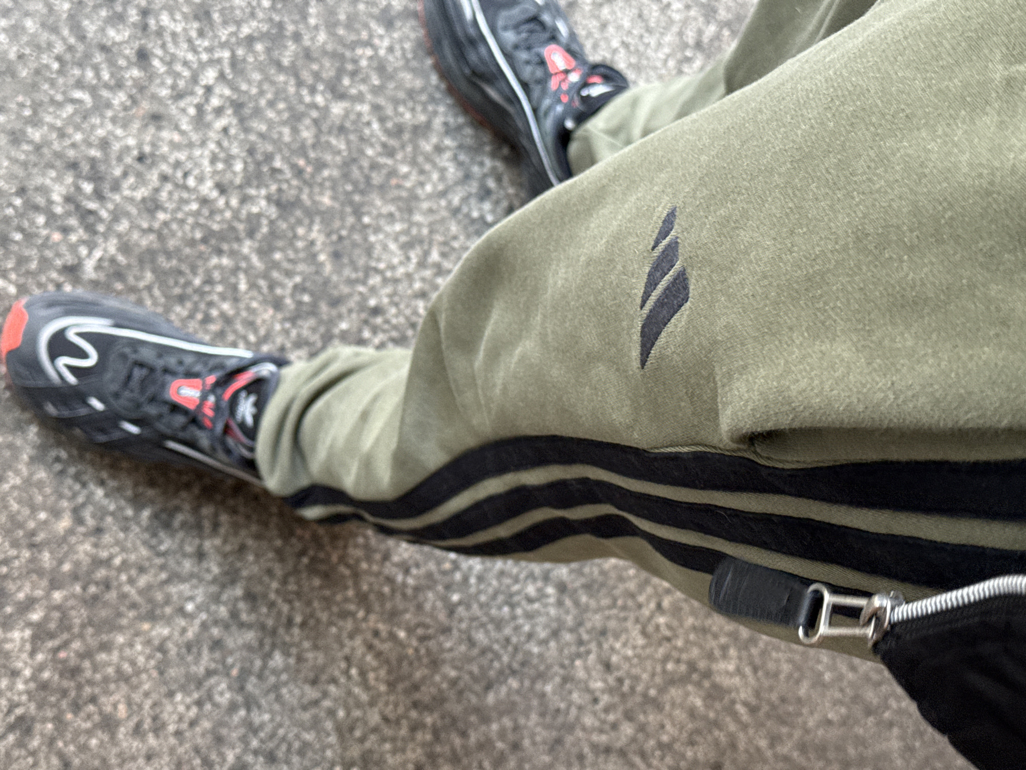 A close-up, top-down shot shows a person standing on a rough gray surface, possibly a street or sidewalk. The person is wearing olive green tracksuit bottoms with black stripes running down the side of the right leg. The tracksuit bottoms feature a small, abstract emblem near the upper thigh. The person wears black trainers with red accents. A black bag with a silver zipper pull is visible near the bottom right of the frame. The location, Stara Zagora, Bulgaria, cannot be verified with the given image information.