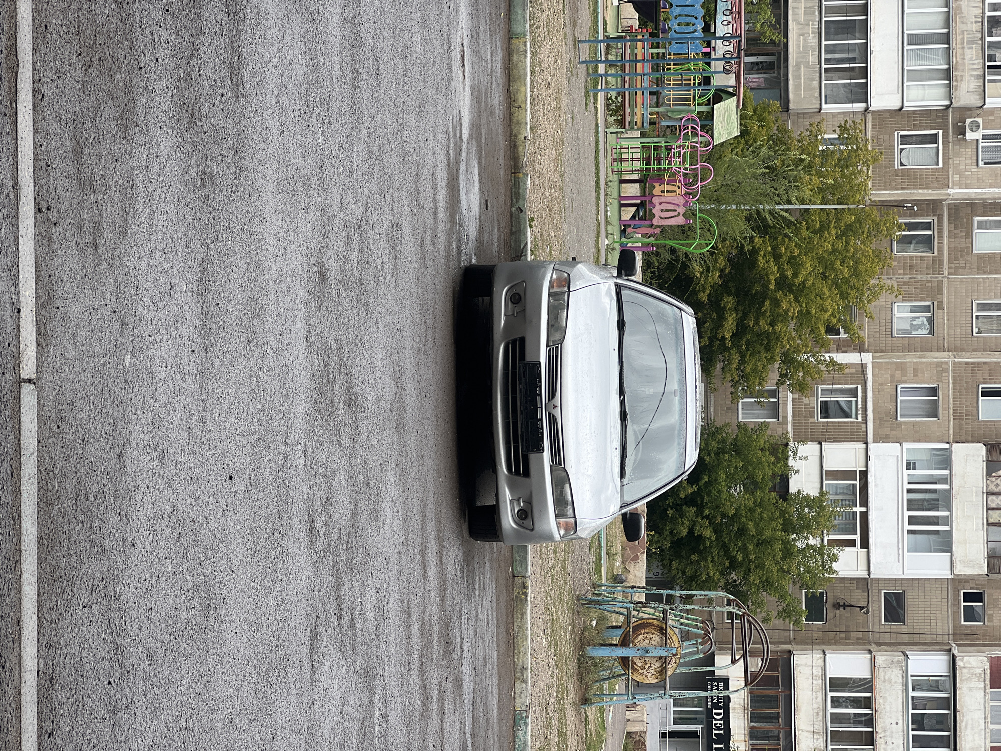 In an outdoor setting in Qarağandı city administration, Kazakhstan, a silver Mitsubishi Lancer is parked on a wet asphalt surface. The car faces the camera, displaying its front bumper, grille, headlights, and windshield, which is speckled with raindrops. Behind the vehicle, a colorful children's playground with swings and slides is visible, along with trees and a multi-story residential building constructed with concrete panels. The ground in the foreground is wet and reflective, indicating recent rainfall. To the right, a rusted metal play structure is partially visible, and a sign for "DEL LI" can be seen on the building. The scene appears to be captured on an overcast day, given the diffused lighting. There are no people visible in the image.