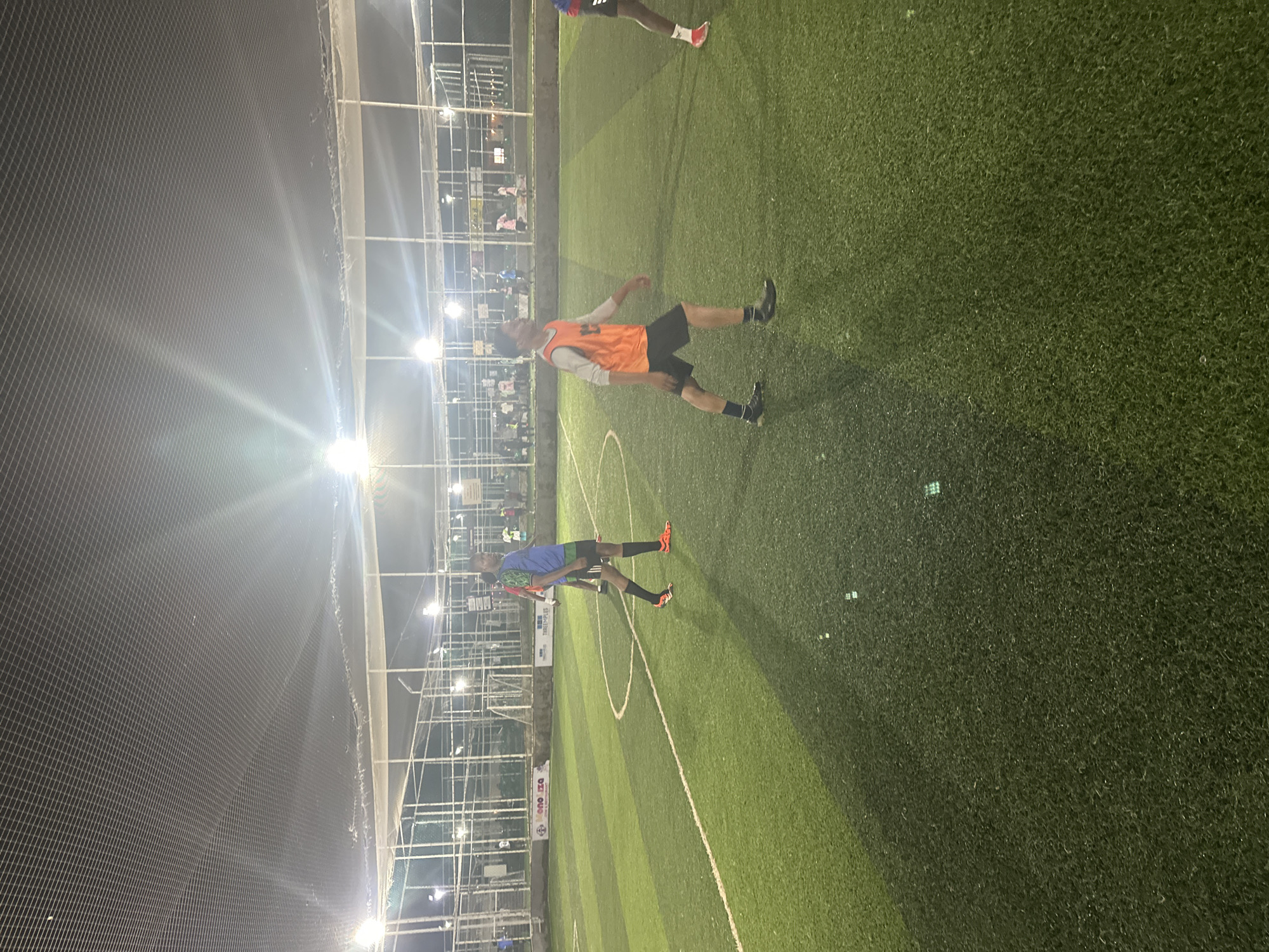 This is a medium-shot, outdoor, night-time photograph of a group of young men playing soccer on an artificial turf field. The field is illuminated by overhead lights, creating strong beams of light and shadows. The field has white lines marking boundaries and a penalty area. In the background, there are white metal fences and netting, suggesting an enclosed sports facility. There are several individuals visible on the field, some actively participating in the game and others observing. One person in the foreground is wearing an orange vest over a grey long-sleeved shirt and black shorts, appearing to be in mid-stride. Another individual to their left is wearing a blue and green soccer jersey and black shorts. The overall environment suggests a recreational soccer match taking place in a sports complex. The image is located in Abuja, Nigeria.