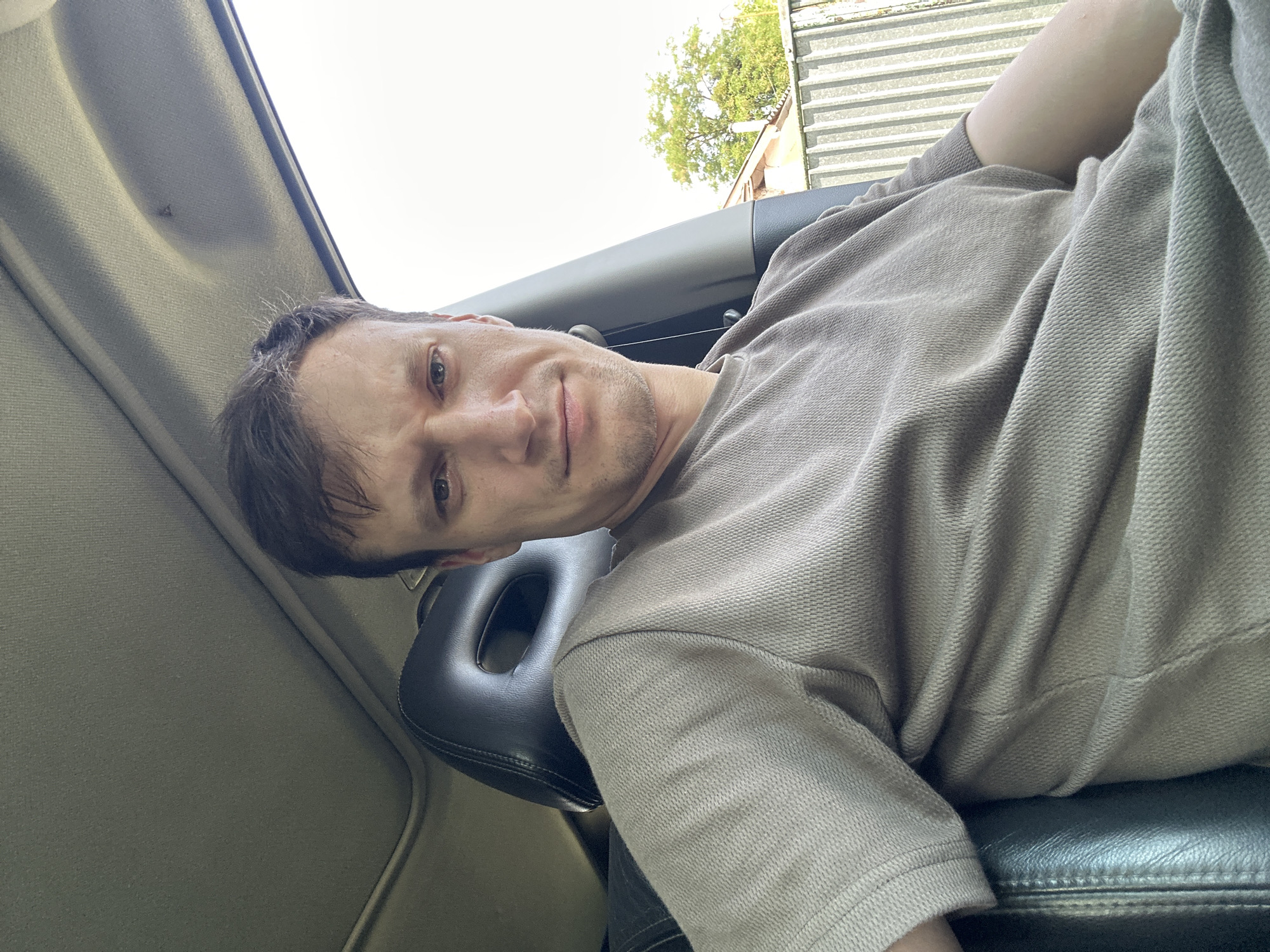 The image shows a man sitting in the driver's seat of a car, looking at the camera. He has short, dark hair and is wearing a light brown, textured t-shirt. The car's interior is visible, including the black leather headrest of the passenger seat and the car's ceiling with a sunroof. Outside the car, a light-colored fence and a tree are visible, suggesting an outdoor location. The man's facial expression is neutral, and he is looking directly into the camera. The lighting indicates it is daytime. The image was taken in Makiv, Ukraine.