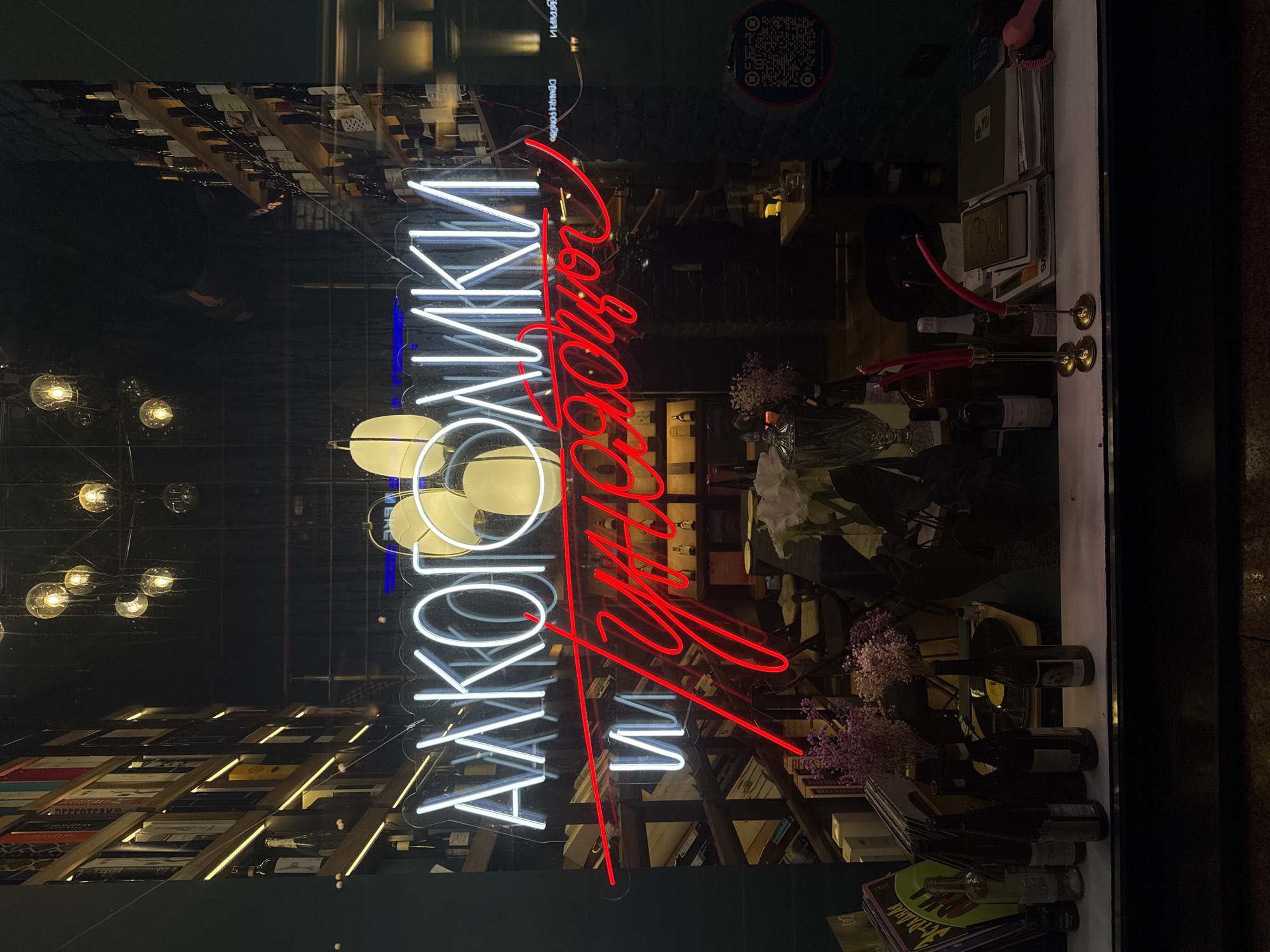 The image shows a shop window display in Moscow, Russia. The words "АЛКОГОЛИКИ" are spelled out in white neon lights, and the words "УДОДОВОЙ" are spelled out in red neon lights. There are bottles of wine, books, and flower arrangements visible inside the store. The reflection in the window shows lights and the interior of another room or building. A QR code is visible in the upper right corner of the window.