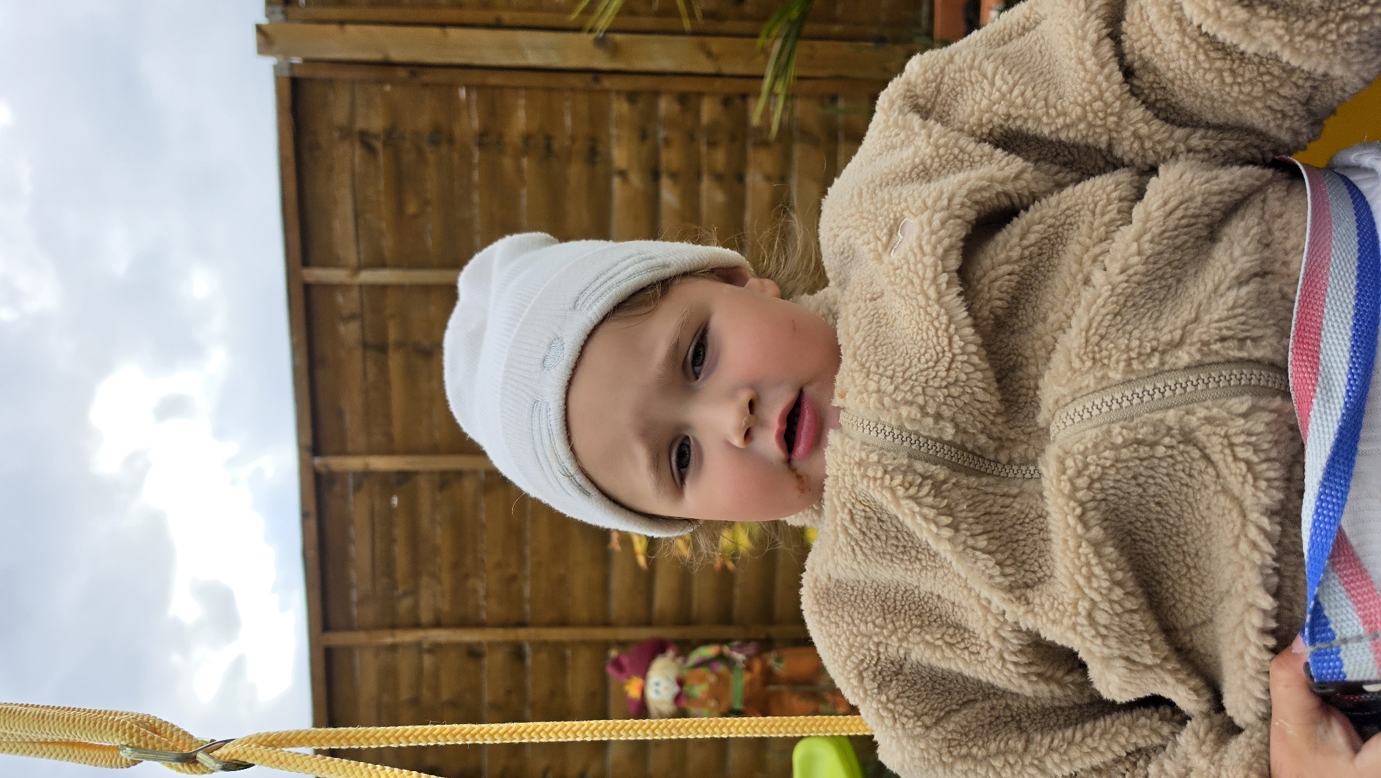 A young child, appearing to be between two and four years old, is prominently featured in the image. The child is wearing a light-colored knit hat and a fuzzy, light-brown jacket. A harness-like strap with red and blue stripes is visible around the child's torso. The child has some smudges near the mouth, possibly from eating.
The background includes a wooden fence or structure, a glimpse of a cloudy sky, and a yellow rope or cable-like element in the foreground. A portion of what seems to be a doll or decorative figure is visible behind the child.
There is no indication in the image or its contents that it is specifically located in London, United Kingdom. Therefore, the location cannot be determined.