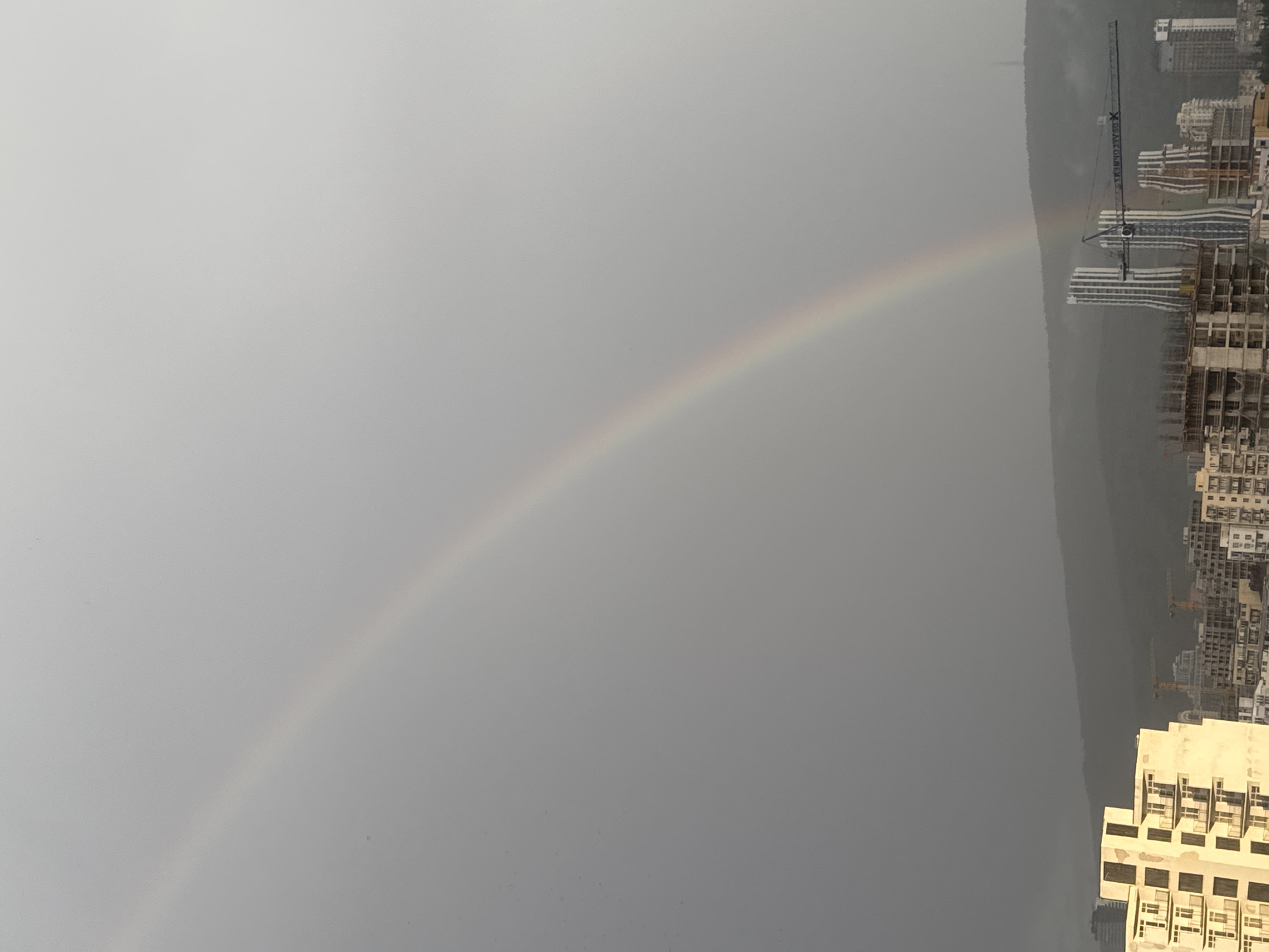 The image shows a view over Tbilisi, Georgia, under a grey sky with a rainbow spanning the horizon. Buildings are concentrated on the right side of the frame, some still under construction, indicated by the presence of cranes. The left side of the image primarily shows the cloudy sky and the arc of the rainbow.