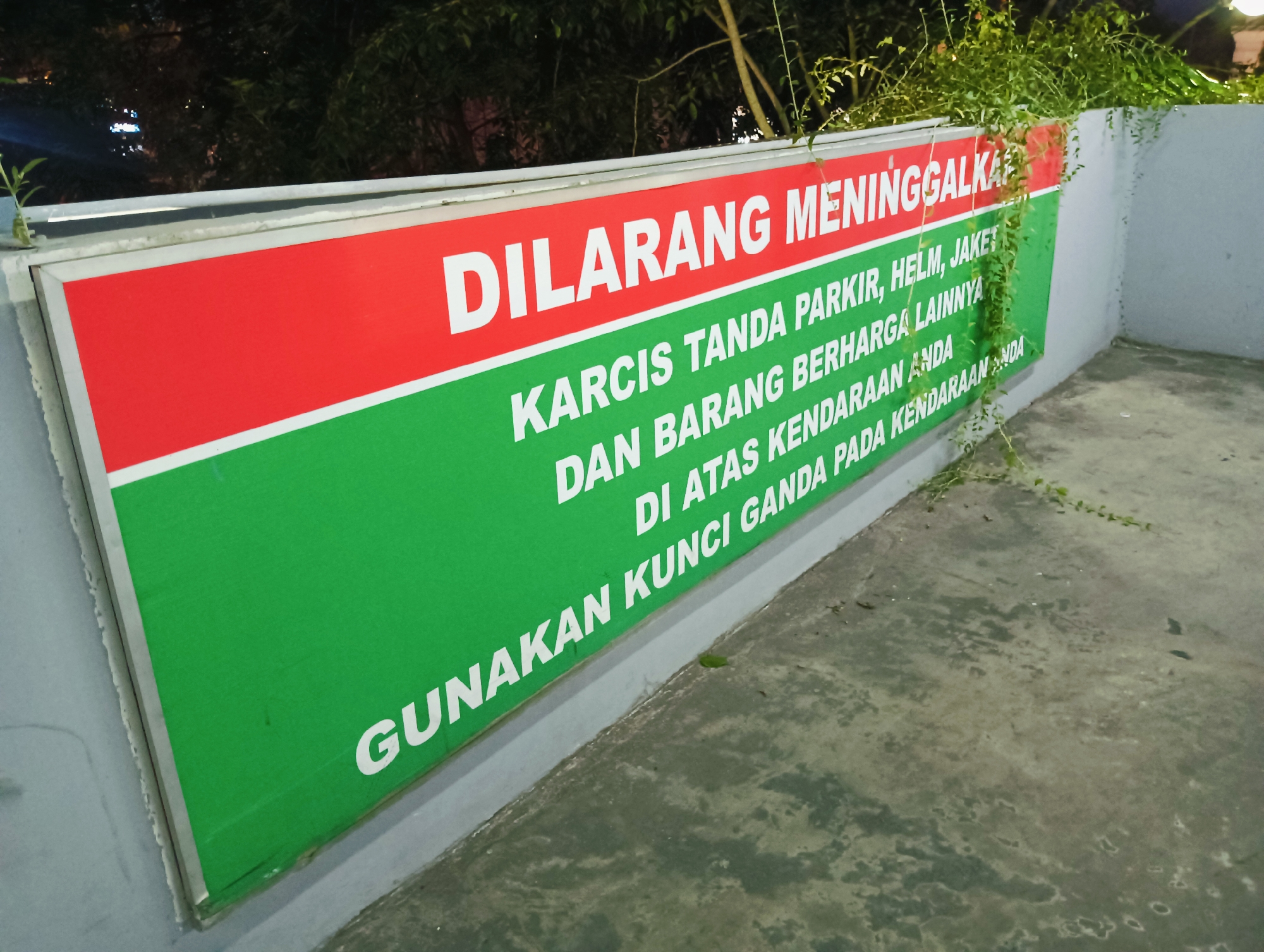 The image shows a sign in Indonesian with the text "DILARANG MENINGGALKAN KARCIS PARKIR, HELM, JAKET DAN BARANG BERHARGA LAINNYA DI ATAS KENDARAAN ANDA. GUNAKAN KUNCI GANDA PADA KENDARAAN ANDA." This translates to "DO NOT LEAVE PARKING TICKETS, HELMETS, JACKETS AND OTHER VALUABLES ON YOUR VEHICLE. USE A DOUBLE LOCK ON YOUR VEHICLE." The sign has a red and green background and is mounted on a grey wall. There are some plants growing on top of and around the sign. The sign is located in what appears to be an outdoor parking area. The ground is concrete and shows signs of wear and tear. The lighting suggests it is either dusk or night. Without further information, it is not possible to definitively determine the exact location within Batam, Indonesia.
