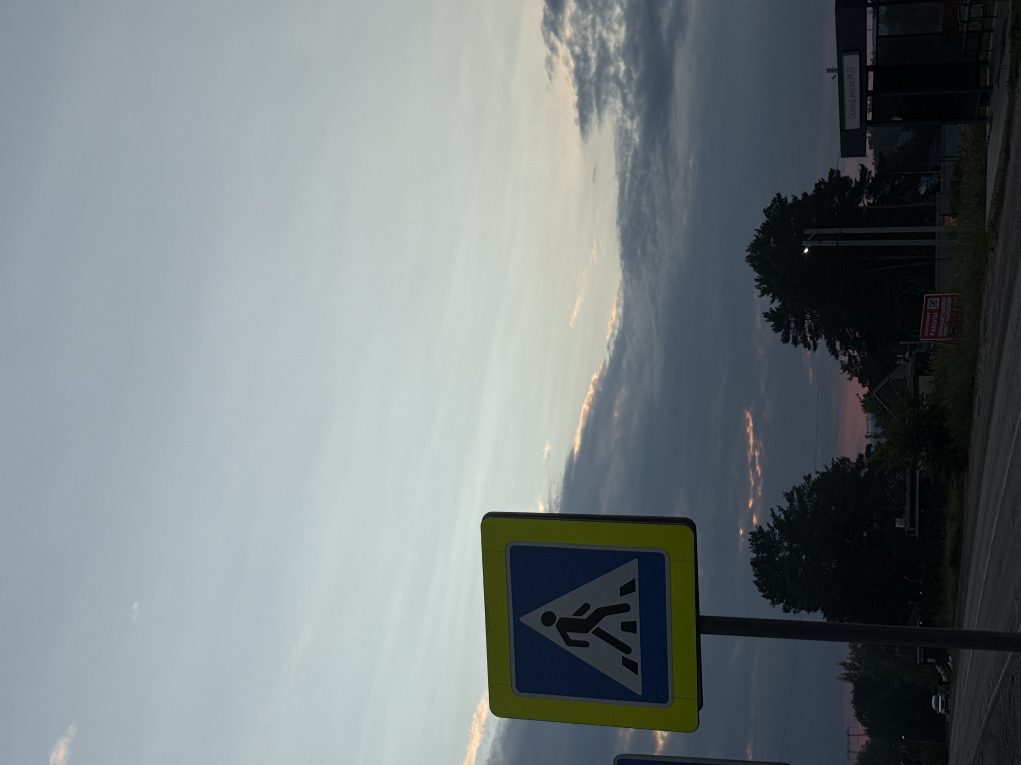 This image depicts a street scene with a prominent pedestrian crossing sign in the foreground. The sign is square with a yellow border and a blue interior, featuring a white silhouette of a person walking across stripes. It is mounted on a grey metal pole. In the background, there is a road, trees, and a bus stop shelter. The sky is partly cloudy, with some dramatic cloud formations illuminated by the setting sun. The location is identified as Chaikovsky, Russia. There are no people visible in the frame. The overall atmosphere suggests a late evening or early morning setting, given the lighting conditions and the colors of the sky.