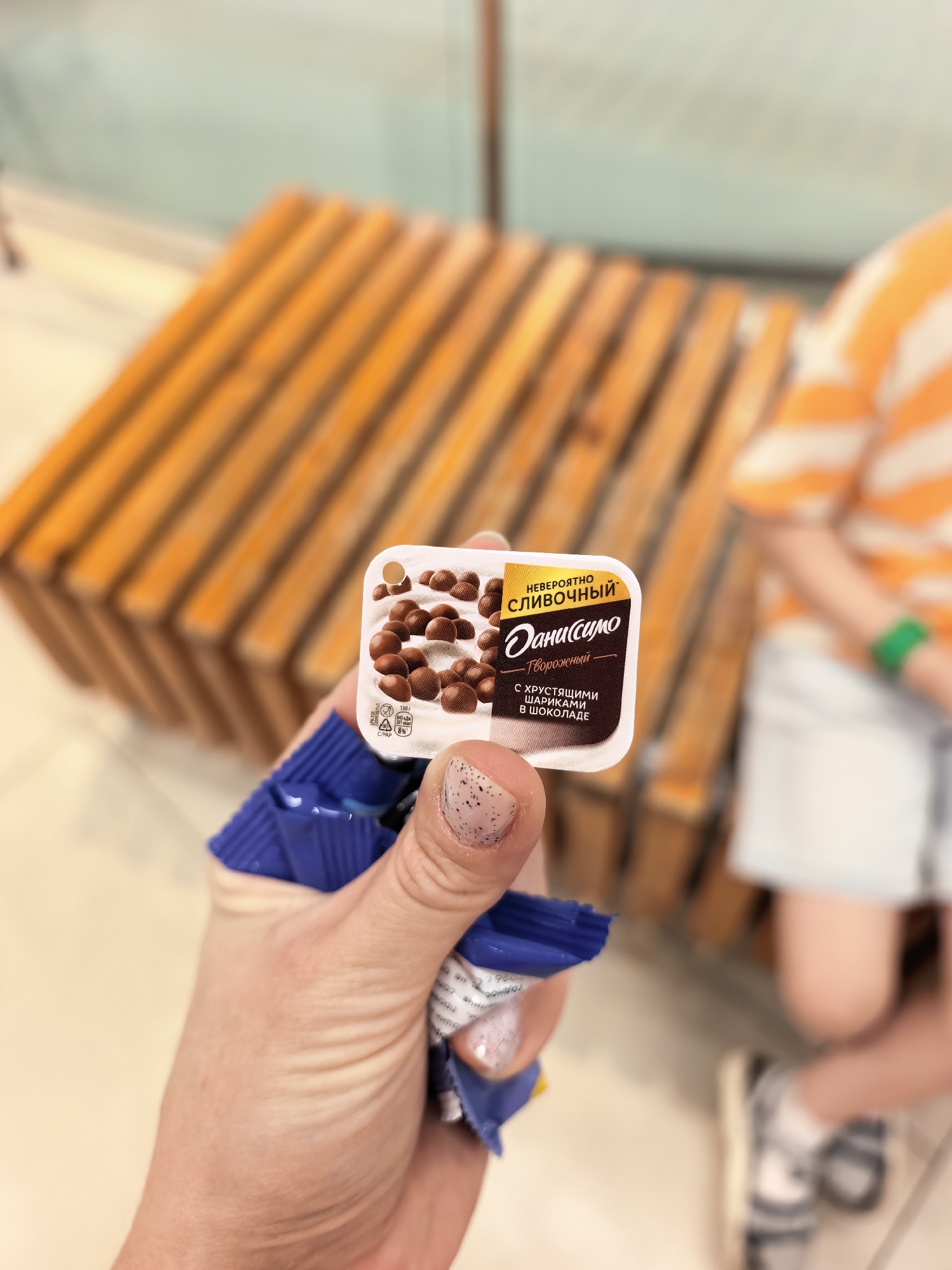 A person's hand holds a small, square, white plastic container of "Danissimo" yogurt with chocolate balls. The container has a lid with a picture of yogurt and chocolate balls, and text in Russian. The container is partially unwrapped from its blue foil packaging. The background is blurred and appears to be an indoor public space with a wooden bench and a child in an orange and white striped shirt and white shorts. The image does not provide any information that would allow for its location in the city of Sterlitamak, Russia.