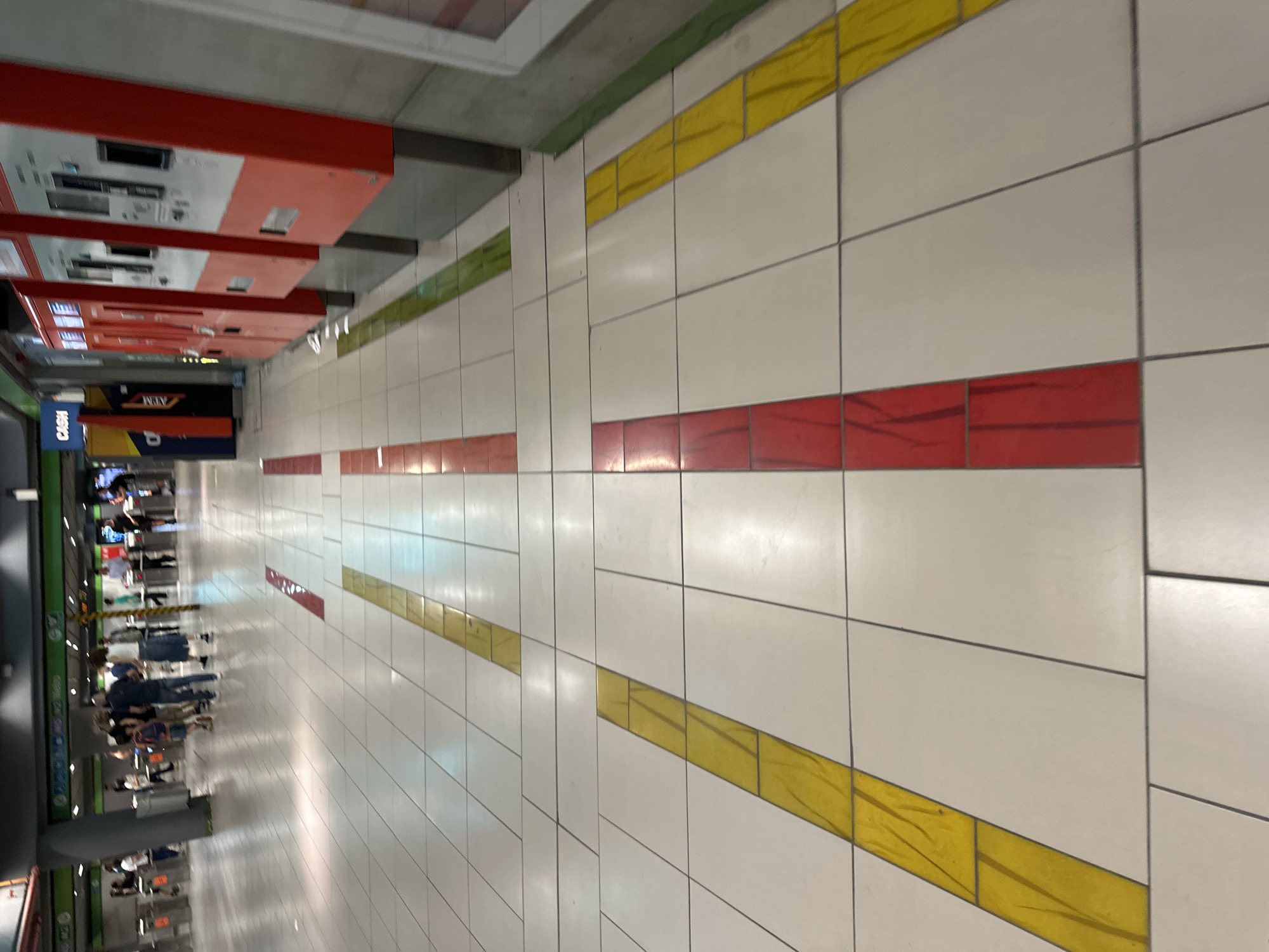 The image depicts a subway station in Milan, Italy, likely located at a station with ticket machines and turnstiles, indicated by the presence of red ticket machines and a row of turnstiles in the background. The floor is tiled with large white tiles and features directional pathways marked by contrasting colored stripes. There are yellow stripes, red stripes, and a green stripe visible. In the foreground, the perspective focuses on these colored stripes on the tiled floor. In the background, there are several people walking through turnstiles and standing around. The lighting appears to be artificial, common for an indoor public transport environment. The scene is clean and orderly, suggesting a well-maintained public space.