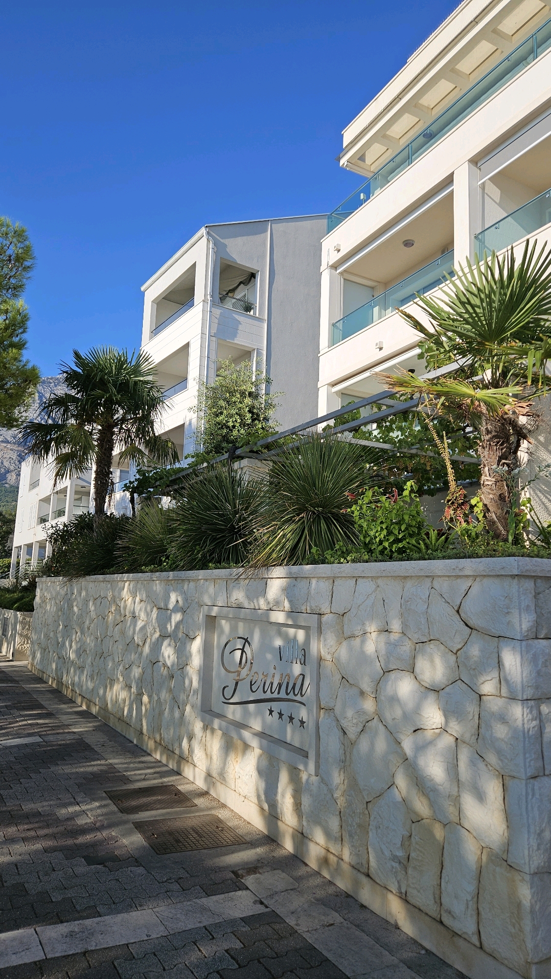 This is a medium-shot, low-angle, eye-level, vertical photograph of "Villa Perina" in Općina Tučepi, Croatia. The building is a modern, white, multi-story structure with balconies. In front of the building, a low, white stone wall runs along a paved walkway. The wall has a sign that reads "Villa Perina" with five stars below it. Lush green foliage, including palm trees and spiky plants, surrounds the building. The sky is clear and blue, and sunlight casts shadows on the walkway. There are no people or animals visible in the image. The overall impression is one of a well-maintained resort or accommodation.