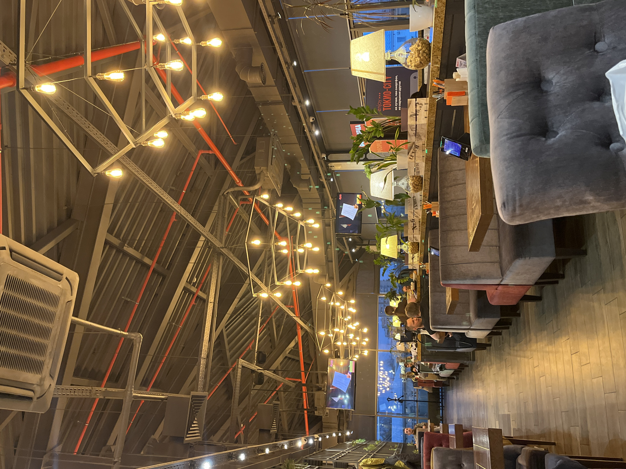 The image captures the interior of a modern restaurant or cafe in Saint Petersburg, Russia, likely during the evening or night, indicated by the warm artificial lighting and the dark exterior visible through large windows.
The scene showcases an industrial-chic aesthetic with a high, exposed ceiling. A complex network of grey metal trusses, red pipes (possibly fire suppression or electrical conduits), and various ventilation components are visible. Numerous bare Edison-style light bulbs are artfully arranged within geometric metal frames, forming large chandeliers that cast a warm, ambient glow throughout the space. Air conditioning units and cable trays are also mounted on the ceiling structure.
Along the right side of the restaurant, a long dining area features upholstered booths or banquettes in shades of grey, green, and possibly red/burgundy, paired with wooden tables. Several patrons are seated, engaged in dining, conversation, or looking at their phones.
Decorations include potted plants arranged along a dividing counter that runs parallel to the seating. Table lamps with fabric shades and glass jars filled with what appear to be peanuts or similar snacks are placed on this counter. Multiple wall-mounted flat-screen televisions display various content, with one clearly showing a document or presentation. The flooring is light-colored wood with a plank pattern.
Visible text on a sign or menu board above the plants includes "ENJOY >>>" and "TOKYO-CITY." Below this, Russian text reads "НЕ ЛУЧШЕ, ЧЕМ СКОРОСТЬ, НО ОЩУЩЕНИЕ ДРУГОЕ," which translates to "NOT BETTER THAN SPEED, BUT A DIFFERENT FEELING."