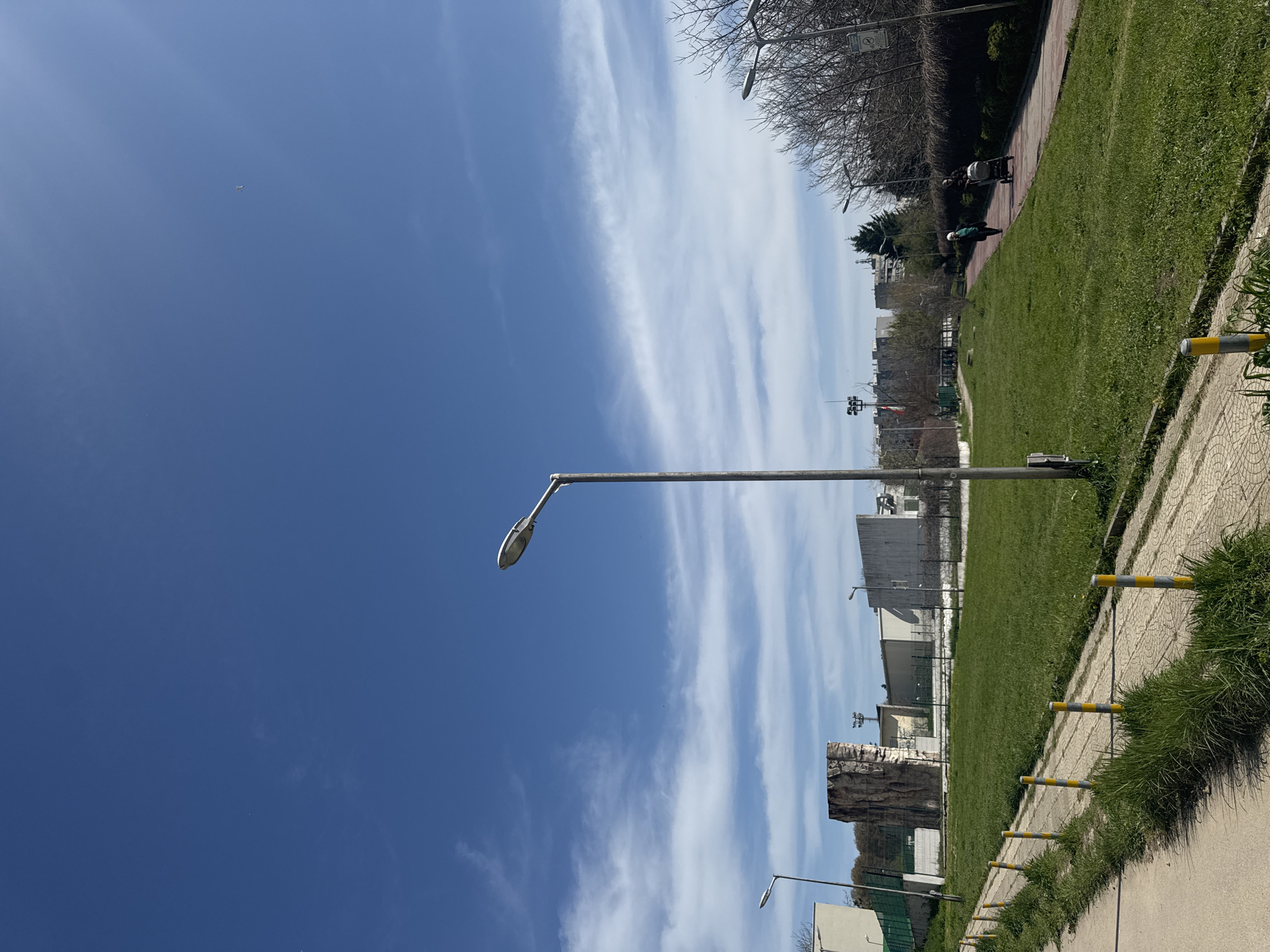 The image depicts a public green space or park under a bright, clear blue sky with thin, wispy white clouds stretching horizontally across. The sun is high, suggesting it is daytime, likely late morning or early afternoon.
In the foreground and mid-ground, a paved pathway runs diagonally from the bottom right towards the upper left, bordered by lush green grass. A series of yellow and grey striped bollards or barriers line the edge of the pathway, separating it from a wider grassy area to the left.
Several streetlights are visible, with one prominent metal lamppost standing centrally, extending into the clear sky. Another streetlight is partially visible in the bottom right, and a few more can be seen further in the background.
On the paved path in the mid-ground, two figures are present. One individual appears to be pushing a light-colored baby stroller, while another person is walking further behind them. They are surrounded by bare-branched trees, indicating either late winter or early spring, though some evergreen trees and shrubs provide greenery in the background.
In the distance, urban structures are visible. These include several buildings, some appearing industrial or utilitarian in design, and a distinctive large, grey artificial rock climbing wall or formation. No legible text is visible on any signs or objects in the image, although a circular sign is faintly discernible on a pole in the upper right background. The overall scene conveys a calm, everyday outdoor environment in Varna, Bulgaria.