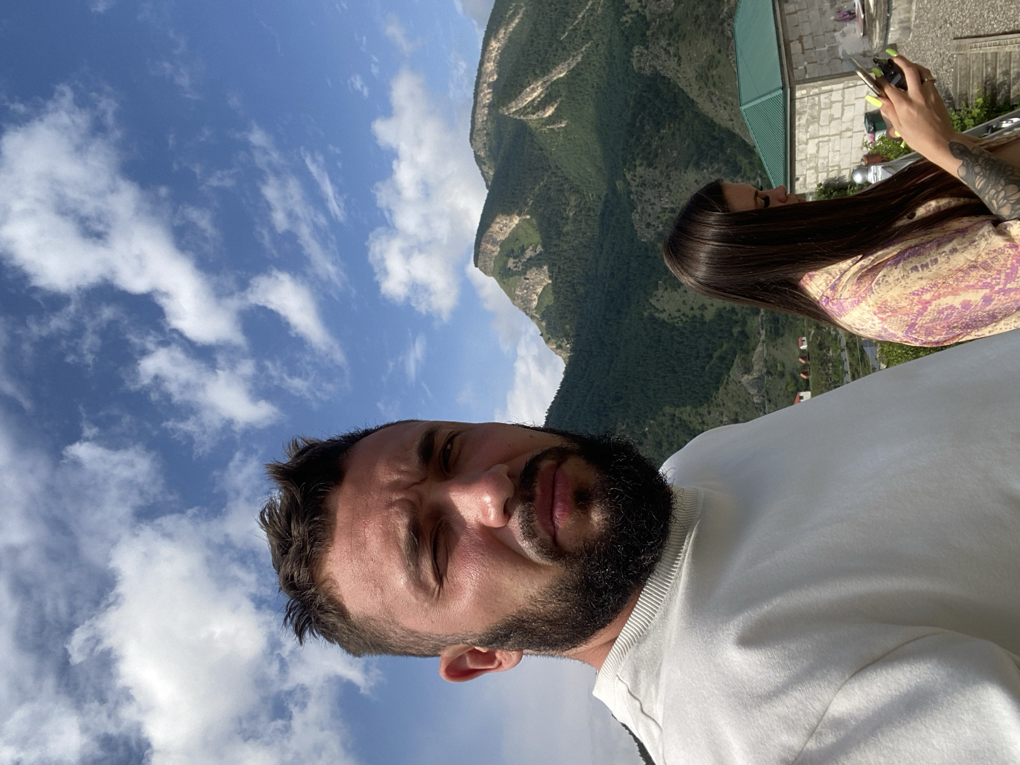 In the foreground, a man with a beard and short dark hair is captured in a close-up selfie. He wears a white crewneck sweatshirt. Behind him, to the right, a woman with long dark hair is visible. She is wearing a patterned shirt and looking down at her phone, which she holds in her hands. Her left arm, adorned with tattoos, is also visible. In the background, a lush green mountain range rises under a bright blue sky filled with white clouds. To the right, a building with a green roof and brick walls is present. The overall scene suggests an outdoor setting, possibly in the mountainous region of Chalda, Russia, given the presence of traditional brick buildings and the mountainous landscape.