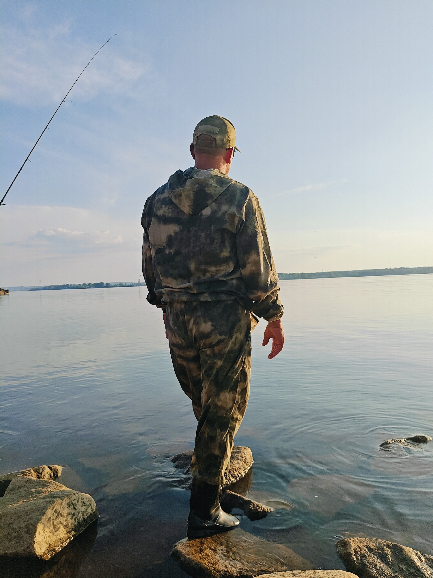 A man wearing camouflage fishing gear and a baseball cap stands on rocks at the edge of a wide, calm river. He is facing away from the camera, looking out at the water. A fishing rod extends upwards from the left side of the frame, with its tip disappearing into the blue sky with scattered clouds. The riverbank on the opposite side is lined with trees and structures, including what appears to be a large industrial crane. The scene suggests a fishing trip on the Kama River in Chaikovsky, Russia.