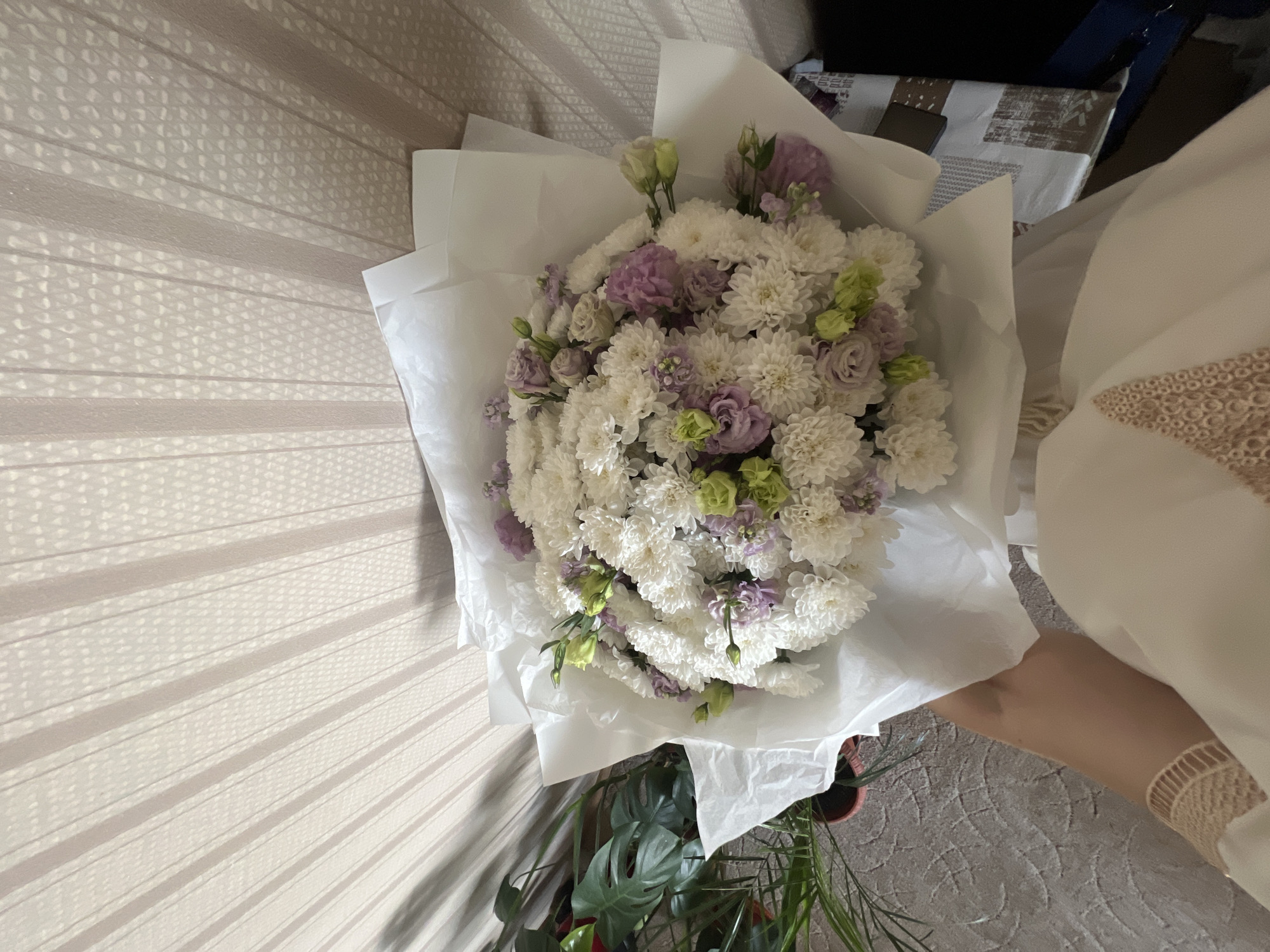 A bouquet of white, purple, and green flowers is held by a person in a white dress. The bouquet is wrapped in white paper and rests against a wall with vertical textured blinds. To the left of the bouquet, a potted green plant with large leaves is visible. The floor is covered with a textured gray carpet. The overall lighting suggests an indoor environment, possibly a home or a flower shop. There is no specific information in the image to indicate the location is in Irkutsk, Russia.