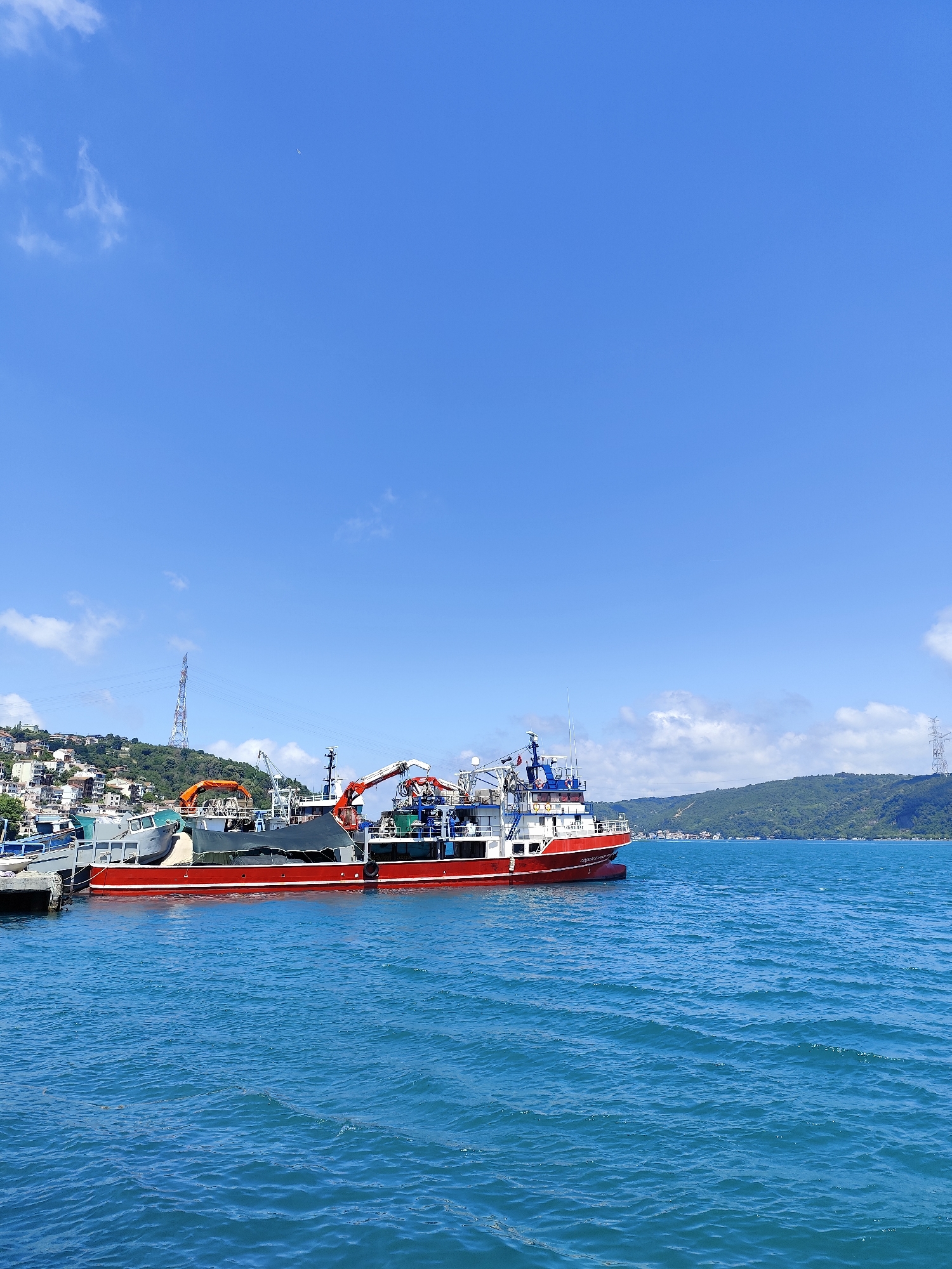An eye-level, full shot captures a body of water with a bright blue sky, dotted with white clouds. In the foreground, the water has gentle waves, reflecting the clear sky. In the mid-ground, a large red and white vessel with cranes is docked, partially obscuring other smaller boats. Behind the boats, a densely populated hillside with buildings and trees rises. In the distance, the sky meets a hazy horizon of hills and scattered buildings. The scene appears to be a harbor or coastal area.