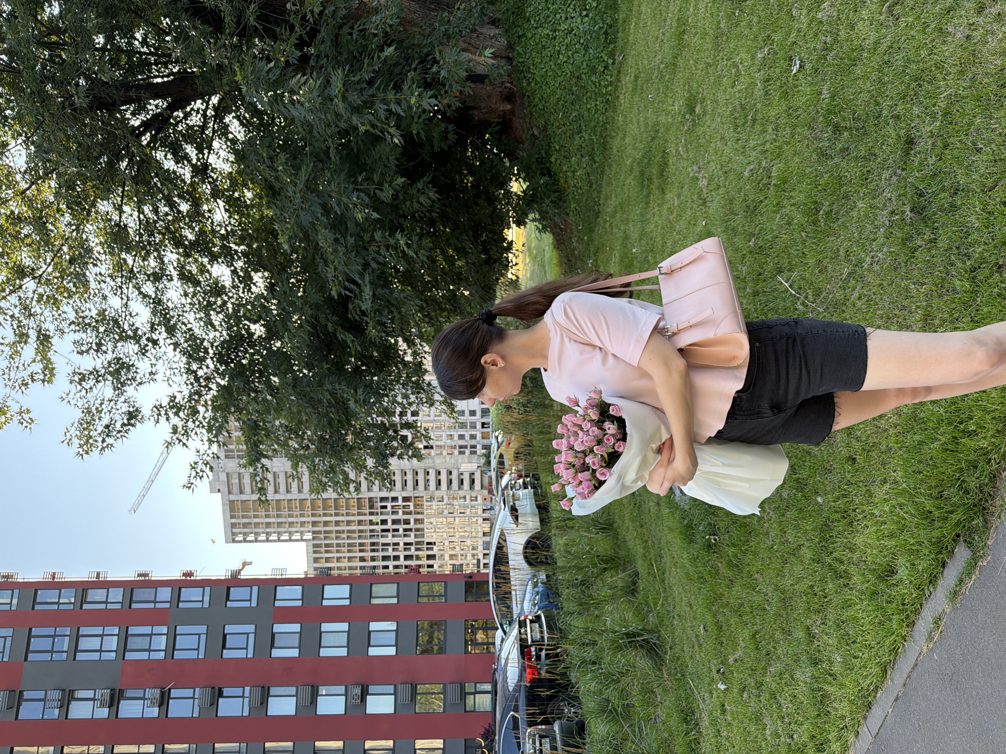 A young woman with dark hair pulled back in a ponytail stands in a grassy area outside of residential buildings in Kyiv, Ukraine. She is wearing a light pink t-shirt and black denim shorts, holding a bouquet of pink roses wrapped in white paper and a light pink purse. The buildings are modern, with red and gray facades, and some are under construction with cranes visible in the background. Cars are parked in the background, and a large, mature tree with green leaves provides shade on the right side of the image. The sky is clear and blue, indicating it is daytime.