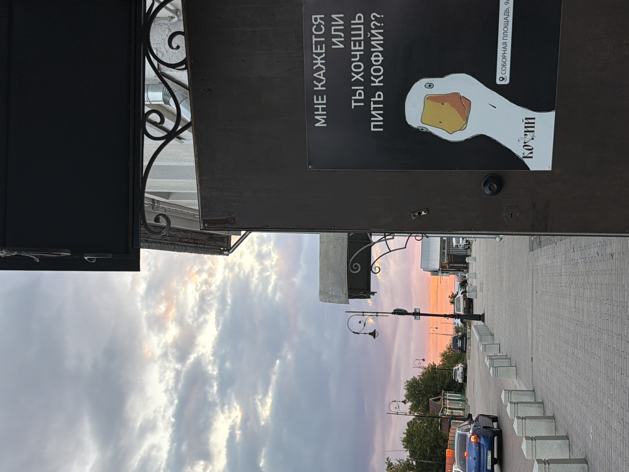 An exterior shot captures a street scene in Arzamas, Russia, with a focus on a dark-colored door featuring a poster. The poster displays text in Russian, "МНЕ КАЖЕТСЯ ИЛИ ТЫ ХОЧЕШЬ ПИТЬ КОФИЙ??", which translates to "It seems to me, or do you want to drink coffee??". Below the text is an illustration of a goose. To the right of the goose illustration, there is a location pin icon followed by text that reads "СОБОРНАЯ ПЛОЩАДЬ, 9А", indicating "Cathedral Square, 9A". The sky above is filled with dramatic clouds tinged with the colors of sunset, transitioning from grey to hues of pink and orange. The street is paved with cobblestones and lined with decorative bollards. Several parked cars are visible along the street, as well as a blue car with a license plate that is partially legible. Trees and buildings can be seen in the background, and streetlights line the pavement.