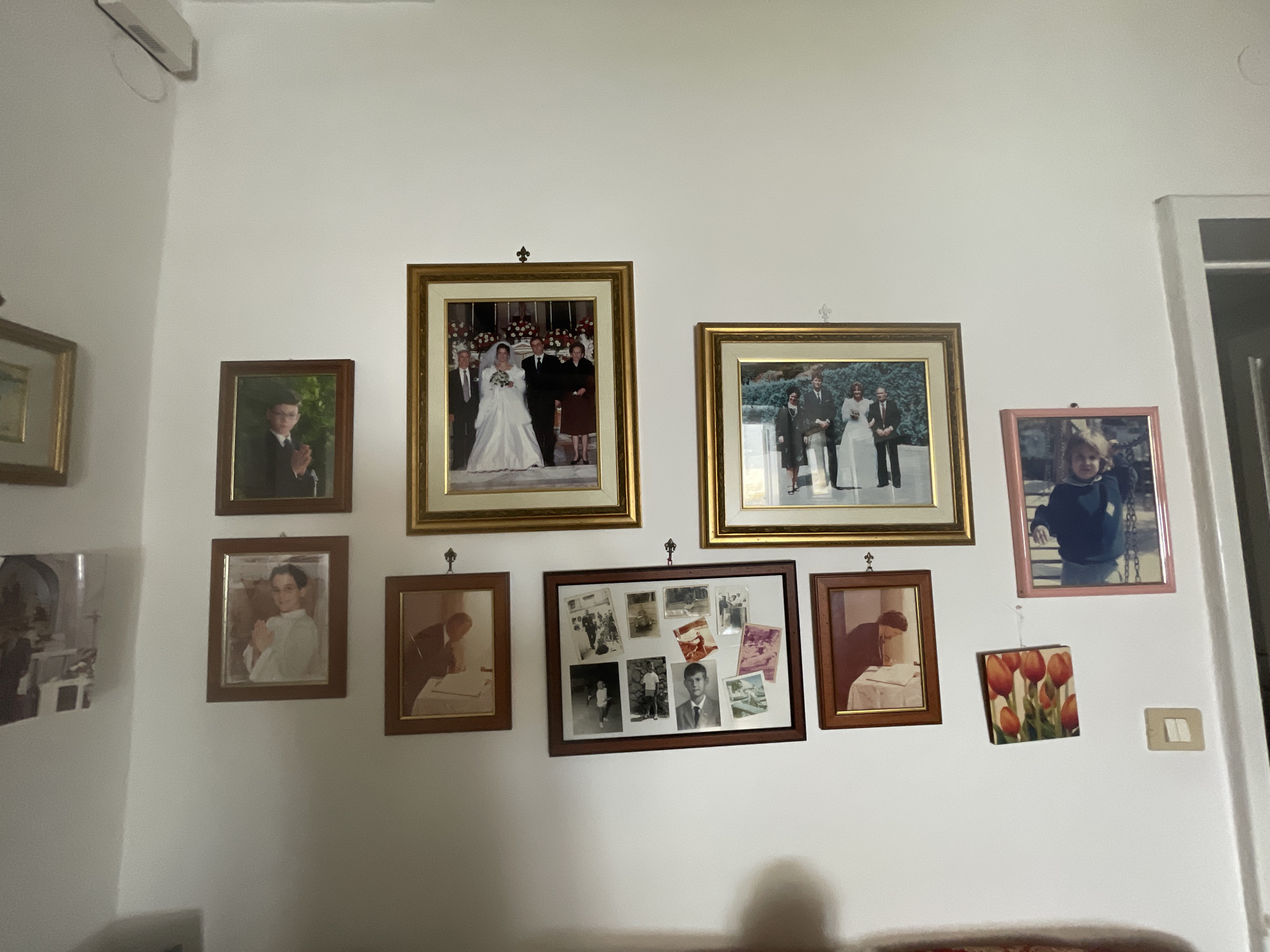 A wall is adorned with numerous framed photographs and a small artwork of tulips. On the left, a large framed picture shows a baptismal ceremony, with a child and several adults. To its right, a smaller portrait features a young boy in a suit. Below this, a framed photograph depicts a man seated at a table, possibly signing a document. Further right, a collage of smaller photographs shows various individuals and scenes. A larger framed photograph of a wedding party, with the bride in a white gown, is prominently displayed above the collage. To the right of the wedding photo, another framed picture shows a group of people, including a bride in a white dress. A pink-framed photograph of a young child is situated in the upper right corner, and a small framed artwork of orange tulips hangs below it. A light switch is visible on the right side of the wall. The wall is painted white, and the scene is lit by artificial light.