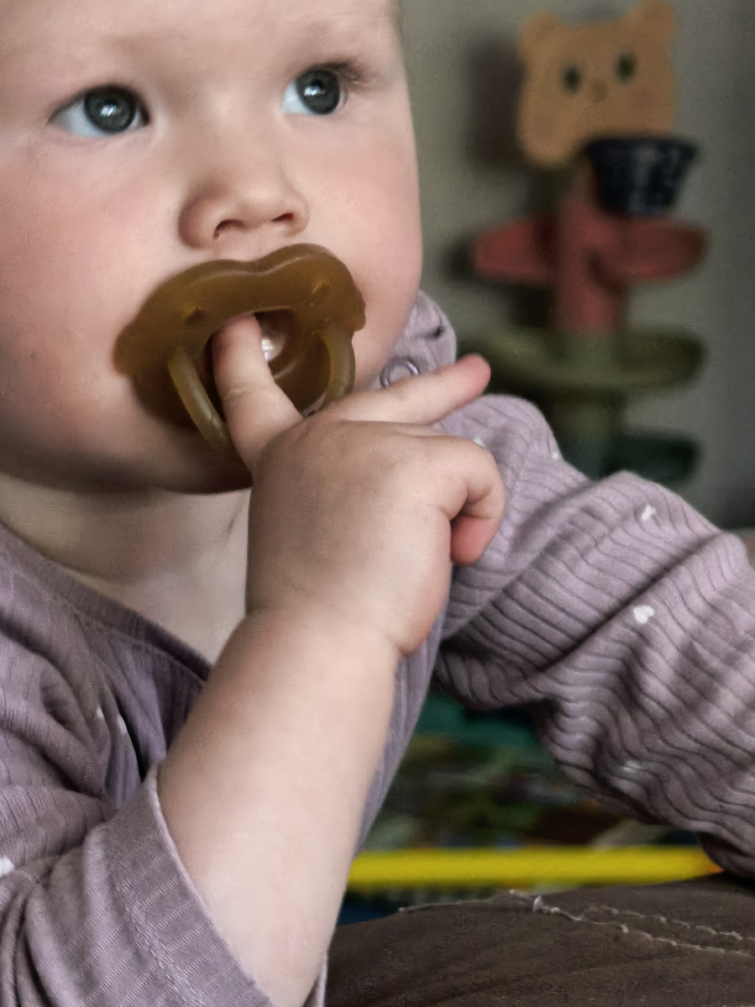 This close-up, softly focused indoor portrait, taken in Sterlitamak, Russia, features a young baby. The baby, with fair skin, chubby cheeks, and striking blue-grey eyes, is positioned to the left of the frame, looking upwards and to the right. A prominent brown or amber-colored natural rubber pacifier is in the baby's mouth. The baby's right hand is raised, with an index finger inserted into the opening of the pacifier, suggesting engagement or comfort-seeking.
The baby wears a light purple or lilac long-sleeved top with a ribbed texture and small white heart patterns.
The background is significantly blurred. To the right of the baby, a distinct but unfocused decorative figure or toy is visible. It appears to be made of wood, featuring a light brown, bear-shaped top, a dark cylindrical section, and a red tiered or cross-shaped middle, resting on a green base. In the lower right, a brown, textured surface, possibly part of a couch or cushion, is present. Hints of yellow and green in the deeper background suggest other items in the room. The lighting is soft and even, indicating an indoor setting. The baby's expression is neutral and focused, absorbed in the tactile experience. No visible text is present in the image.