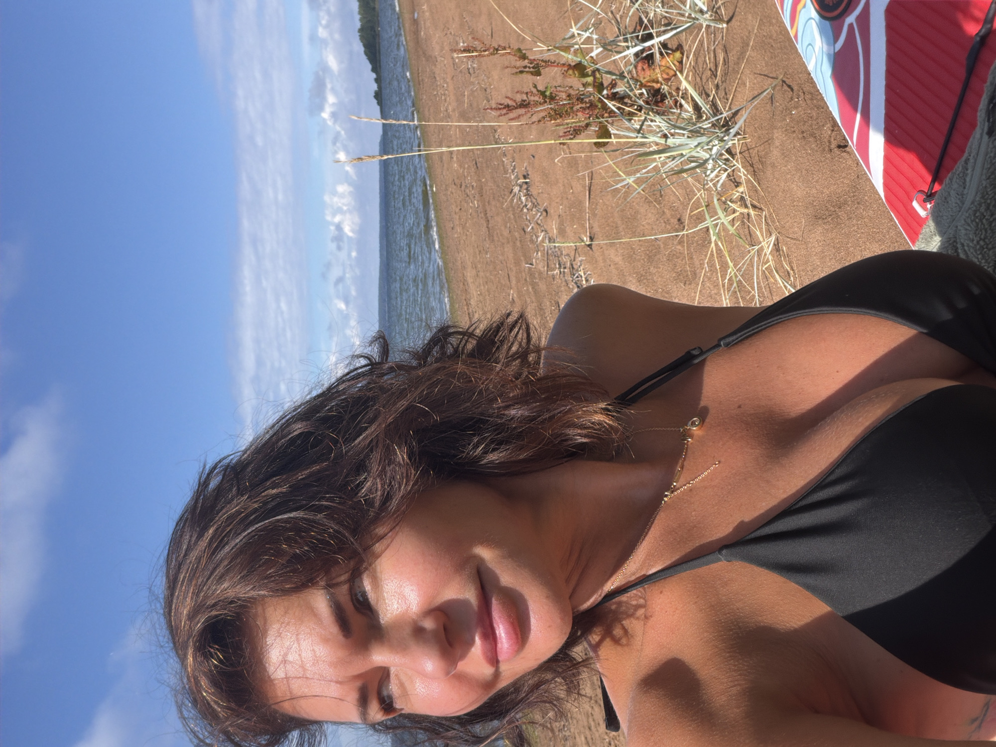 A woman with brown hair and fair skin is taking a selfie on a sandy beach. She is wearing a black bikini top and a gold necklace. The background shows a body of water, a sandy shore, and a blue sky with clouds. The woman is looking to her left with a slight smile. The image appears to be taken on a sunny day. There is no information available about the specific location in the city of Vyazy, Russia.
