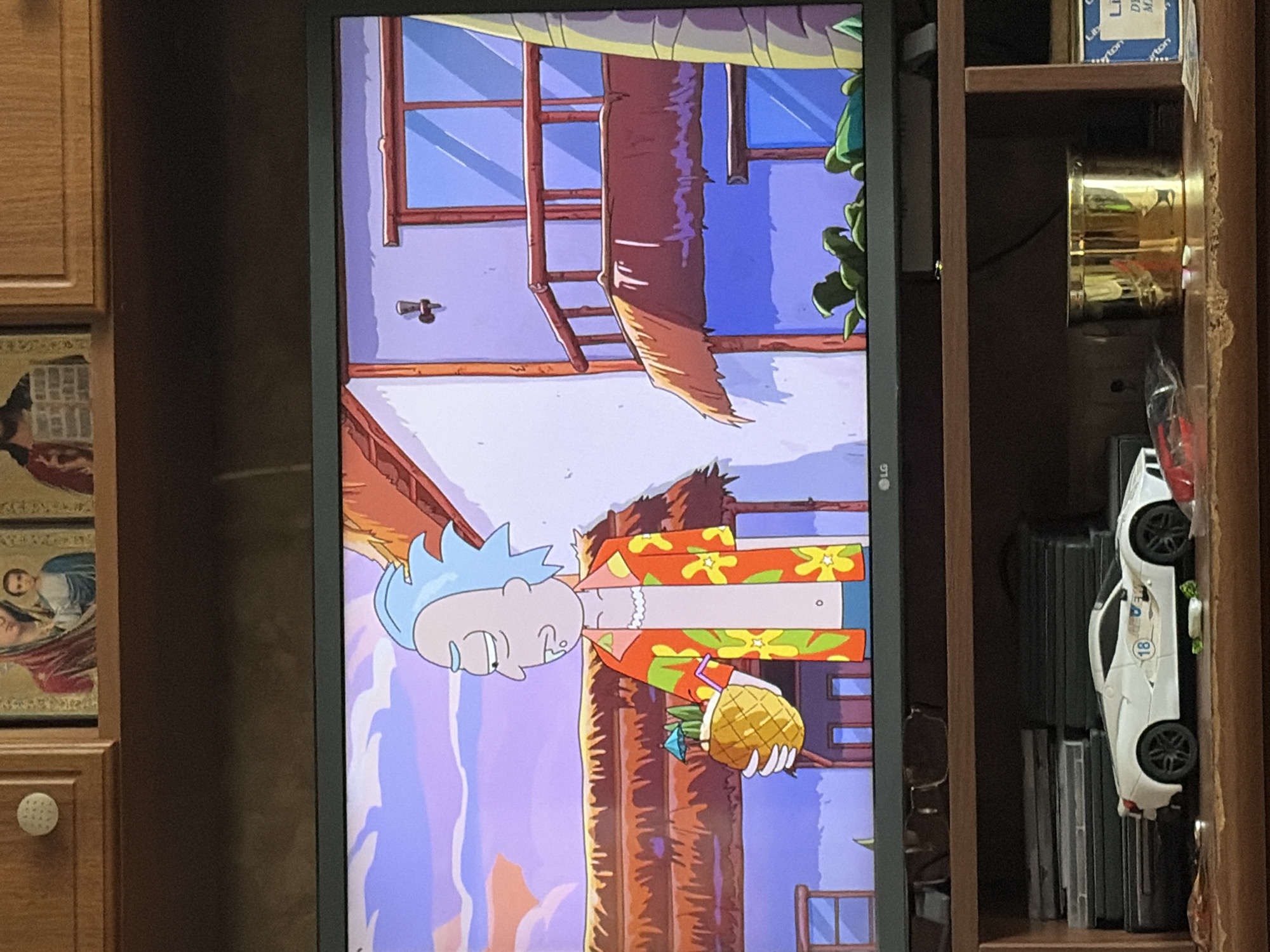 The image shows a television screen displaying an animated scene from Rick and Morty. Rick Sanchez, with his signature blue spiky hair and a Hawaiian shirt, is holding a pineapple cocktail. He is standing in front of a thatched-roof building with a balcony. The background features a purple and pink sky with clouds. Below the television, a wooden shelf holds a white toy car, stacks of DVDs, and a gold-colored container. Religious icons are visible on the cabinet above the television. The scene appears to be set in a tropical or beach-like environment.