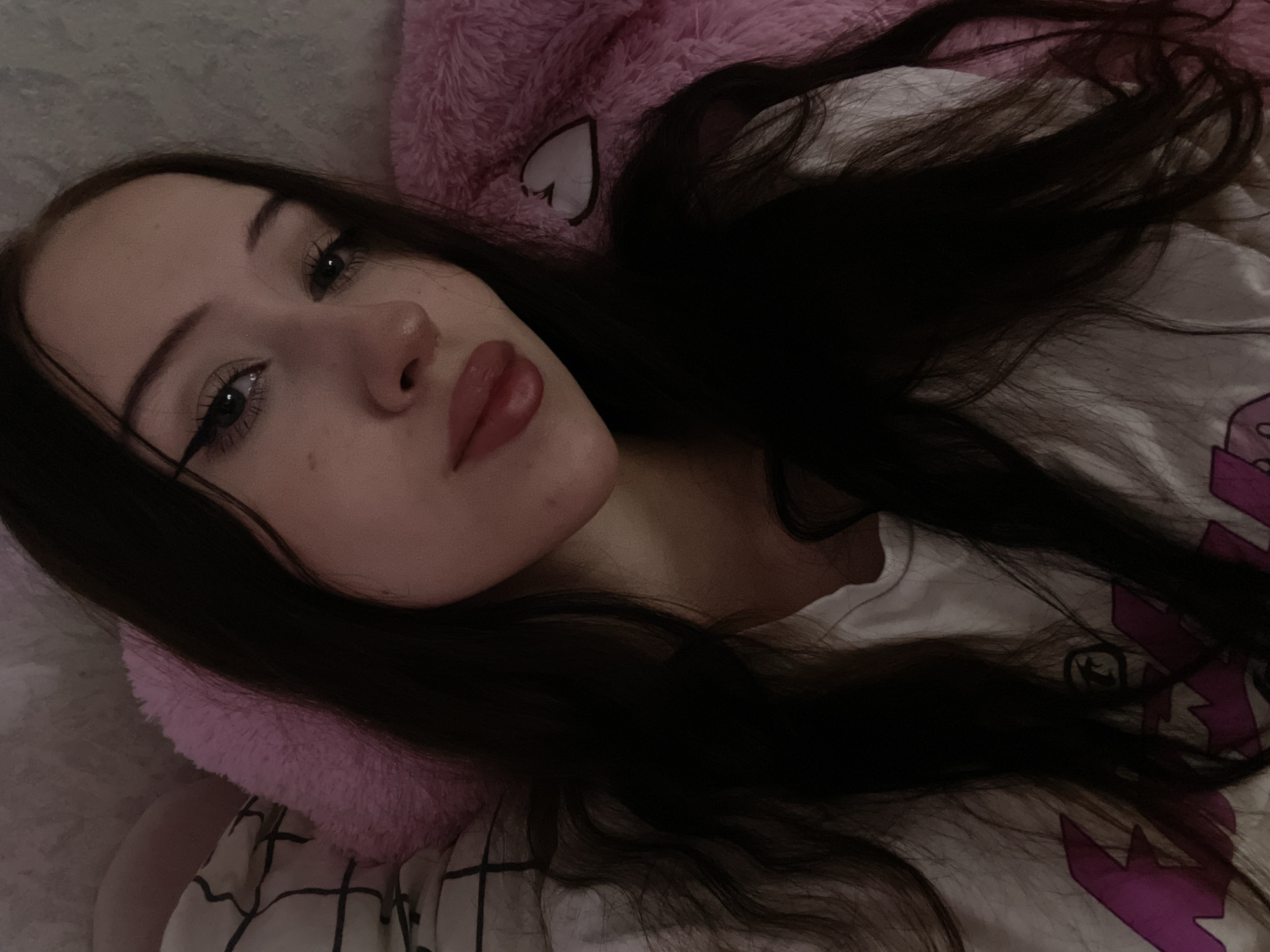 A young person with long, dark hair lies on a pink fuzzy blanket. They are wearing a white t-shirt with a graphic design. Their face is turned towards the camera, and they have eye makeup on. The background appears to be a bedroom setting. There is no indication of location in Votkinsk, Russia.