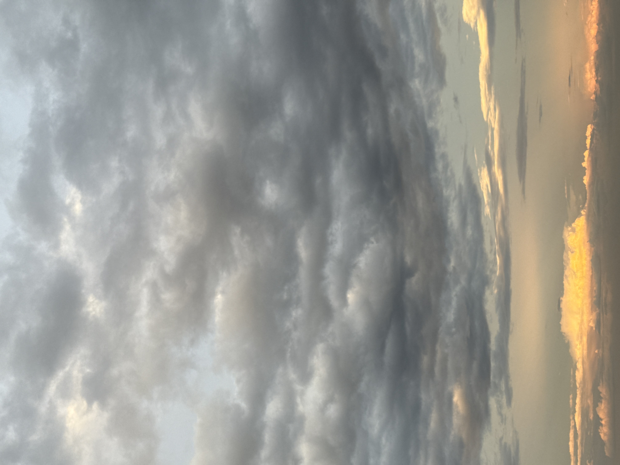 A photograph of the sky shows a cloudscape, with the left side of the frame dominated by thick, textured, gray clouds. These clouds appear heavy and dense, with subtle variations in tone and some areas catching the light. Towards the right side of the image, the clouds are thinner, lighter in color, and tinged with hues of yellow and orange. These brighter clouds suggest the presence of sunlight, potentially indicating either sunrise or sunset. The overall impression is of a sky filled with dramatic weather conditions, possibly a storm approaching or receding. The location in the city of Unknown, Unknown.