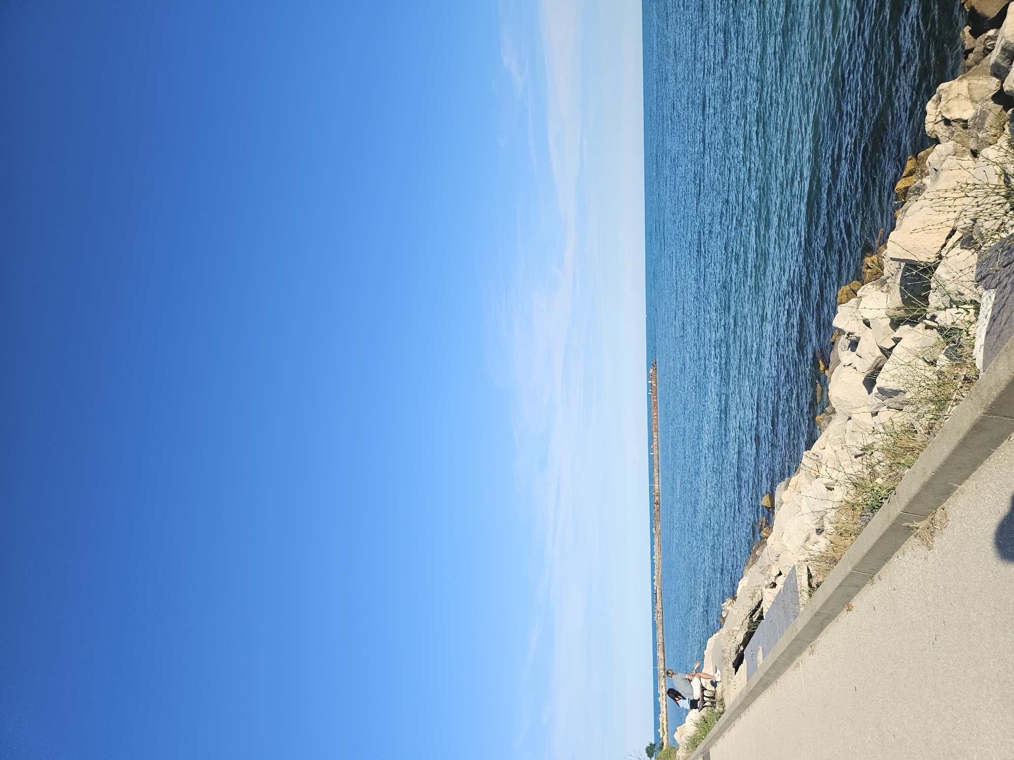 A bright, clear day is depicted, with a vast expanse of blue sky dominating the left side of the image. The sky transitions to a lighter blue near the horizon, where it meets the calm, turquoise water. The ocean stretches out to the right, its surface rippling gently. Along the right edge of the water, a rocky shore is visible, composed of large, light-colored stones. Some sparse, dry grass grows between the rocks. A paved walkway runs parallel to the shore, separating the rocks from the viewer. In the distance, a long pier or breakwater extends out into the water, dotted with small structures or lights. On the walkway, two people are sitting. One person, wearing dark clothing, appears to be leaning back, while the other, wearing a light-colored shirt and dark pants, is seated beside them. The overall scene suggests a peaceful day at the coast, likely in the late morning or early afternoon given the bright sunlight and clear sky. There is no visible text in the image.