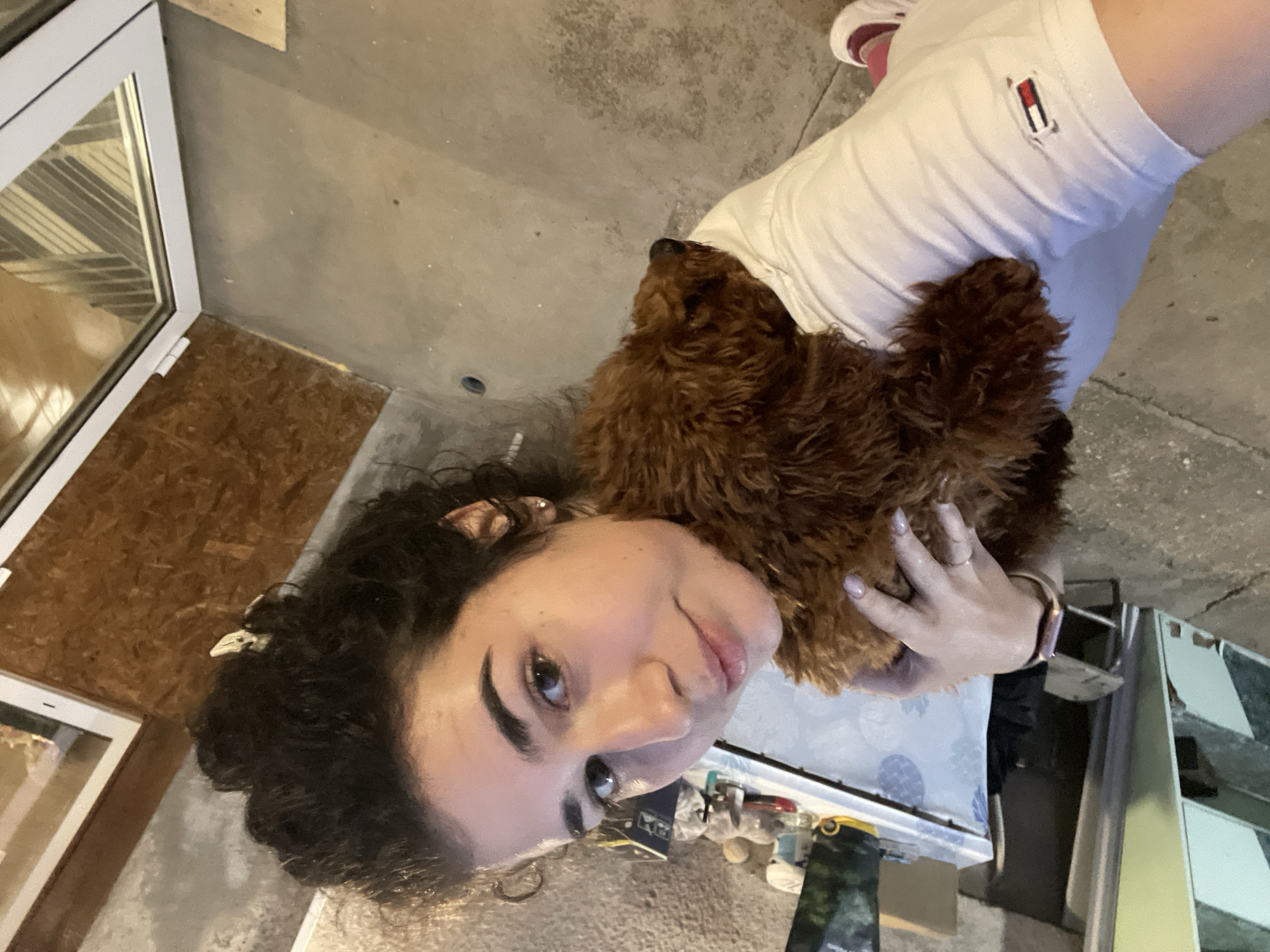 A woman with curly dark hair and fair skin is holding a brown poodle. She is wearing a white shirt with a Tommy Hilfiger logo. A watch is on her left wrist. The scene appears to be indoors. Behind the woman and to the left is a window frame. The floor is concrete and there is some wood paneling on a wall. Without additional information, it is not possible to determine a specific location within Sofia, Bulgaria.