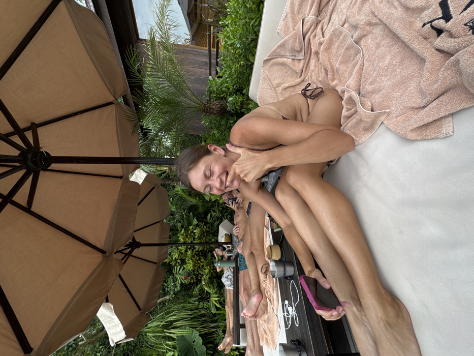 An outdoor scene in Canggu, Indonesia, depicts four individuals relaxing on white cushioned lounge chairs under large beige patio umbrellas. In the foreground, a female with light brown hair, wearing a dark-patterned bikini, is seated. Her eyes are closed, her mouth is open in a wide smile, and her right index finger touches her lower lip. A pink smartphone is held in her left hand, resting on her left thigh. A light pink towel with a dark emblem is draped over the lounge chair to her right. To her left, three other individuals are reclining on separate lounge chairs. Two individuals are partially visible, lying prone with bare legs and feet, wearing dark swim trunks. The third, further back, is also lying prone, partially obscured, with a device held to their face. Dark water bottles, metal tumblers, a wooden bowl, and white wired headphones are present on dark low tables situated between the lounge chairs. The background features lush tropical vegetation, including large-leafed plants and palm trees. A dark-roofed wooden building is partially visible in the upper right background. The sky appears overcast.
