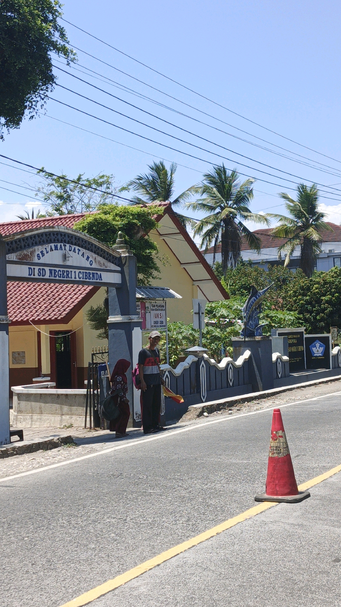 The image depicts the entrance to SD Negeri 1 Cibenda in Cibenda, Indonesia, on a sunny day. The school's entrance gate has a sign that reads "SELAMAT DATANG DI SD NEGERI 1 CIBENDA." Two people are standing on the sidewalk in front of the school. One is a woman wearing a hijab and a patterned dress, carrying a large dark bag. The other is a man wearing a t-shirt with red, white, and black stripes, dark pants, a hat, and carrying a yellow and red flag. A red traffic cone is placed on the road in the foreground. The school building has a red tiled roof and yellow walls, with lush green trees and palm trees surrounding it. Overhead, power lines stretch across the clear blue sky.
