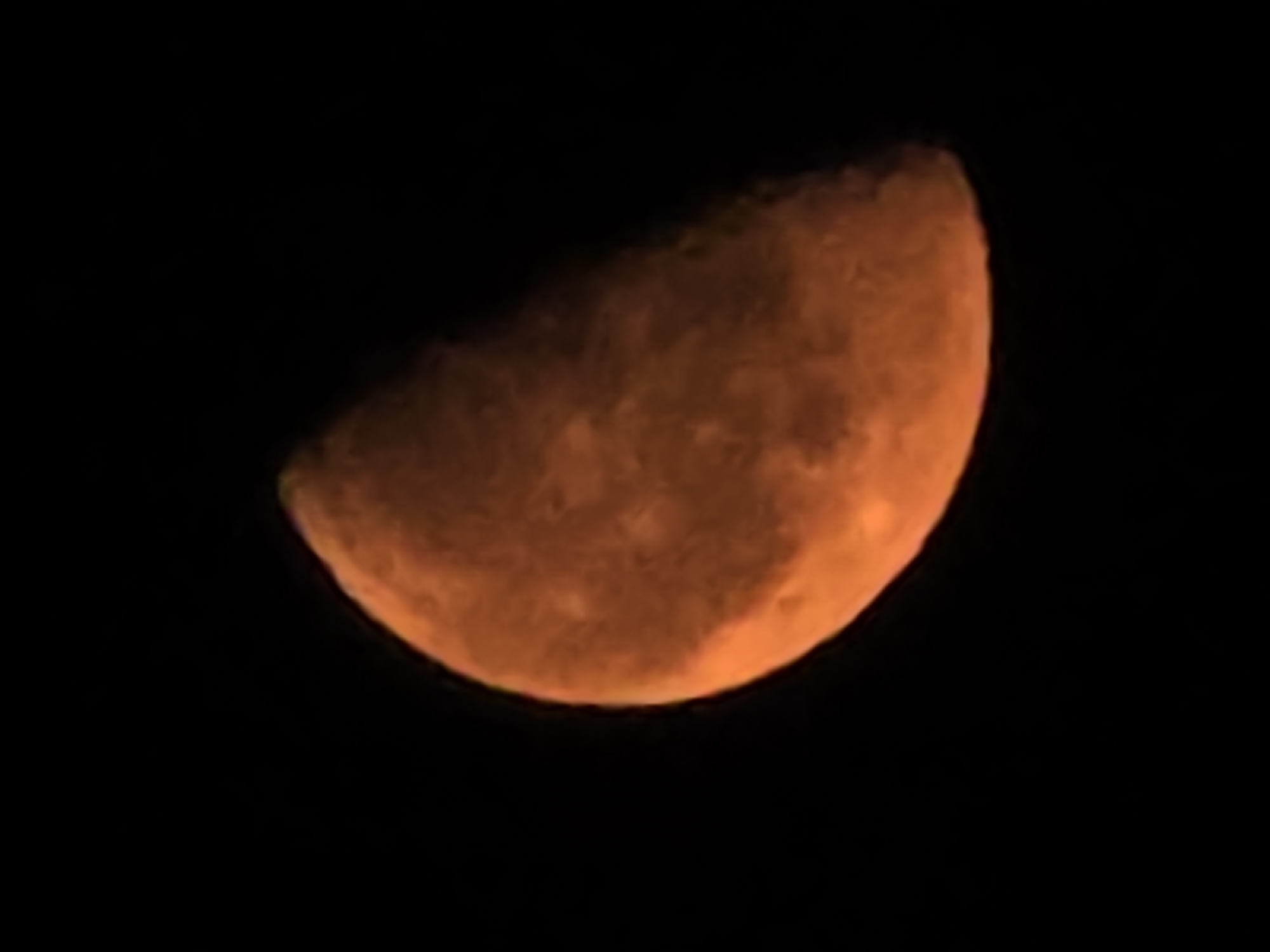 An astronomical photograph depicts a partial view of the moon against a black sky. The moon, illuminated by an unseen light source, appears to be roughly half-covered, with the illuminated portion taking on a reddish-orange hue. The surface of the moon shows some textured detail, suggesting craters or other geological features, though the resolution is not high enough for precise identification. The image is devoid of any terrestrial elements or context, offering no information about its specific location within Yambol, Bulgaria, or any other geographical setting.