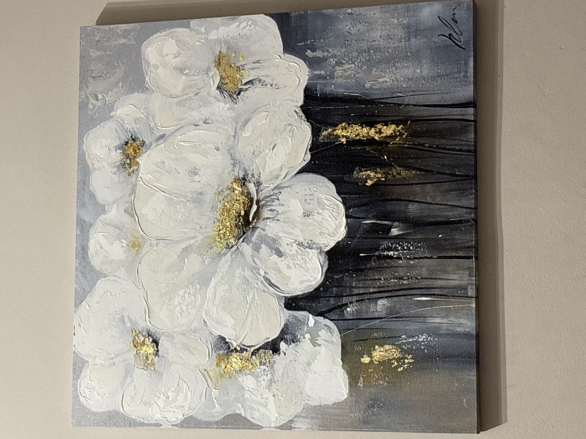 The image displays a rectangular canvas painting, approximately square, mounted on a light-colored, flat wall. The artwork features an abstract composition of white and off-white flowers, rendered with significant impasto texture, suggesting depth and volume. Gold leaf accents form the centers of the flowers and are scattered throughout the lower portion of the painting, which transitions from a light grey and muted blue background in the upper half to dark, vertical strokes resembling stems or a dark vase in the lower half. A dark, cursive signature is visible in the bottom right corner of the canvas. The painting casts a minor shadow on the wall beneath its bottom edge, indicating its physical protrusion. This scene is located in the city of Sofia, Bulgaria.