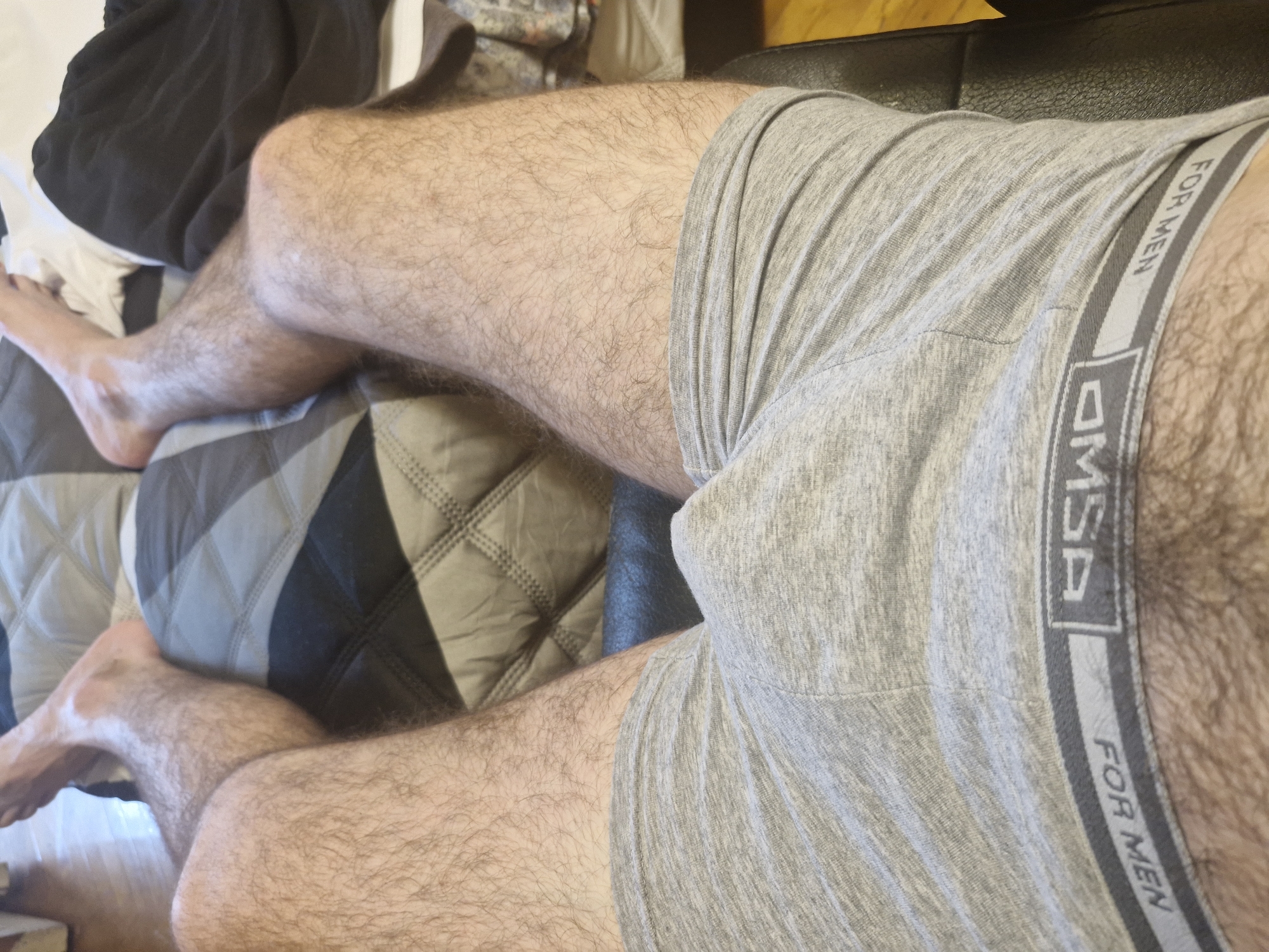 This image shows a close-up view of a person's legs and torso, clad in grey boxer briefs. The person is lying down on a surface that appears to be a bed or sofa, with a patterned blanket or comforter visible beneath them. A dark fabric, possibly clothing or bedding, is also present in the upper left corner of the frame. The lighting suggests it could be daytime, with natural light illuminating the scene. The boxer briefs have a prominent waistband with the text "FOR MEN" and a logo that includes the letters "MSP". The person's legs are covered in hair, and the overall setting appears to be indoors, possibly a bedroom. The image is cropped tightly, focusing on the lower body and underwear. No specific emotions are conveyed, and the activity seems to be resting. The location context of Lyubertsy, Russia, does not add any visual cues to the image itself.