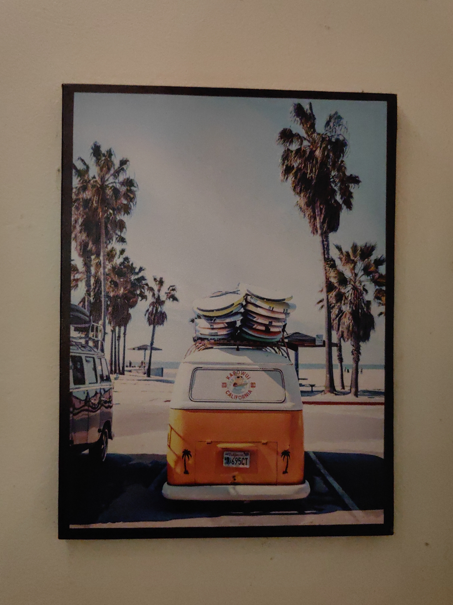 The image is a framed photograph of a beach scene with two Volkswagen vans. The main van, painted orange and white, is in the foreground with surfboards strapped to its roof. The license plate reads "695CT". The other van, partially visible to the left, is dark in color. Palm trees line the beach, and the sky is bright. "Ka'oulu California" is written on the van. The location is not in Cao Lộc, Vietnam, but likely California based on the text on the vehicle.