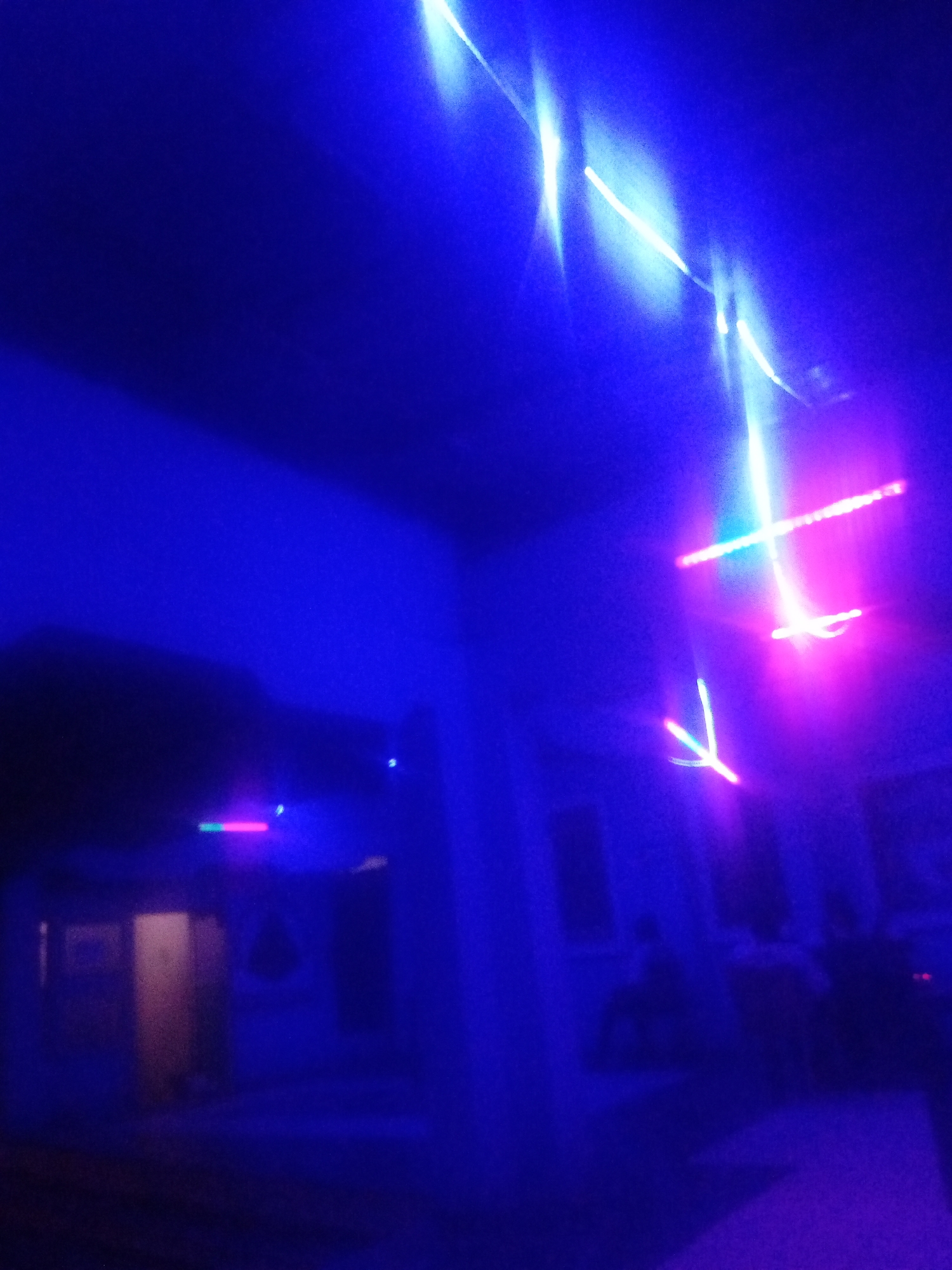 The image captures an indoor scene bathed in deep blue and purple lighting, likely from a nightclub or entertainment venue. Streaks of bright white and pink light crisscross the upper portion of the frame, suggesting moving lights or laser projections. In the background, blurry figures can be discerned, appearing to be people gathered in a space with doorways and walls. The overall ambiance is dark and electric, with the vibrant artificial lights dominating the visual field. There are no discernible landmarks or specific details that would pinpoint a location within a city.