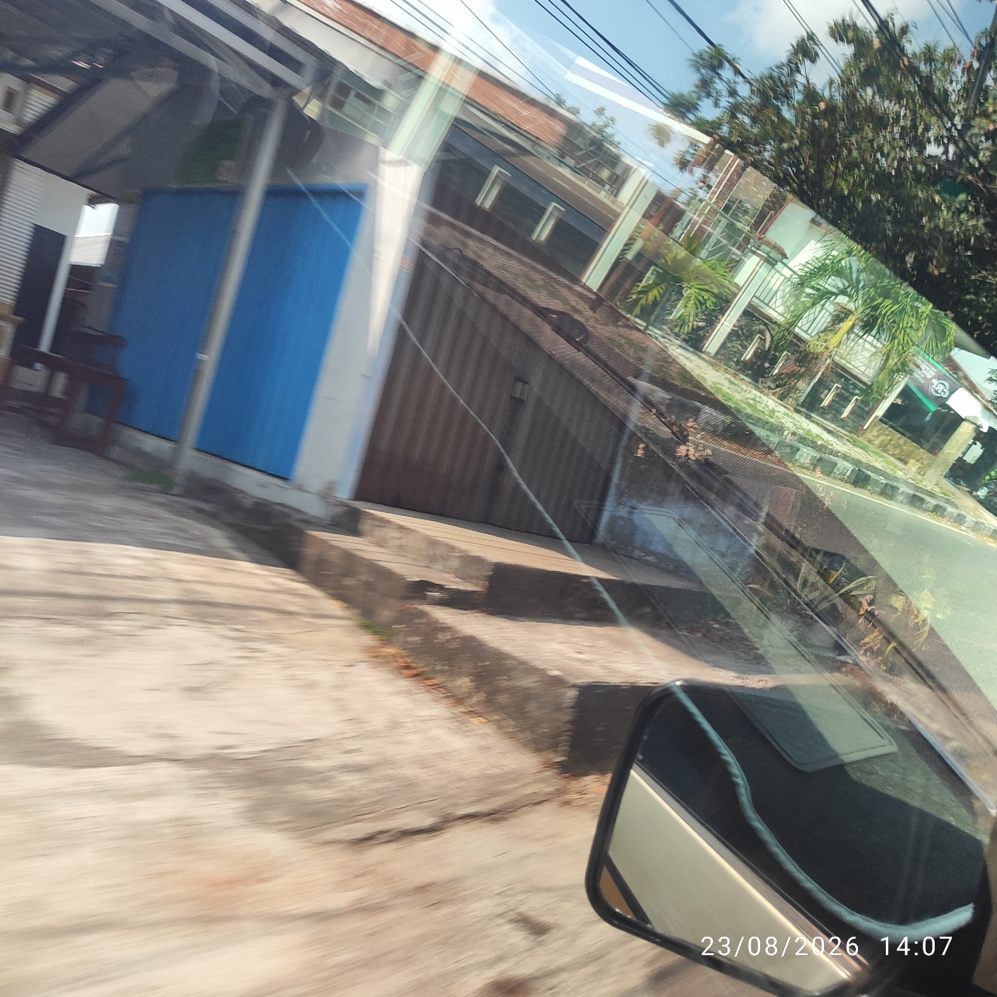 The image is a photograph taken from inside a vehicle, possibly from the driver's perspective, looking out through the windshield and side mirror. The timestamp "23/08/2026 14:07" is visible in the bottom right corner, suggesting a future date. The scene captures a street view in Tanjung Pendam, Indonesia, characterized by buildings with corrugated metal and tiled roofs, a prominent blue door, and a gray concrete pathway. Overhead, electrical wires are visible against a bright sky with some clouds. In the foreground, the vehicle's side mirror is reflected in the glass, showing the interior of the car and the driver's seat. The overall impression is that of a snapshot of a passing moment on a street in a tropical or subtropical urban environment.