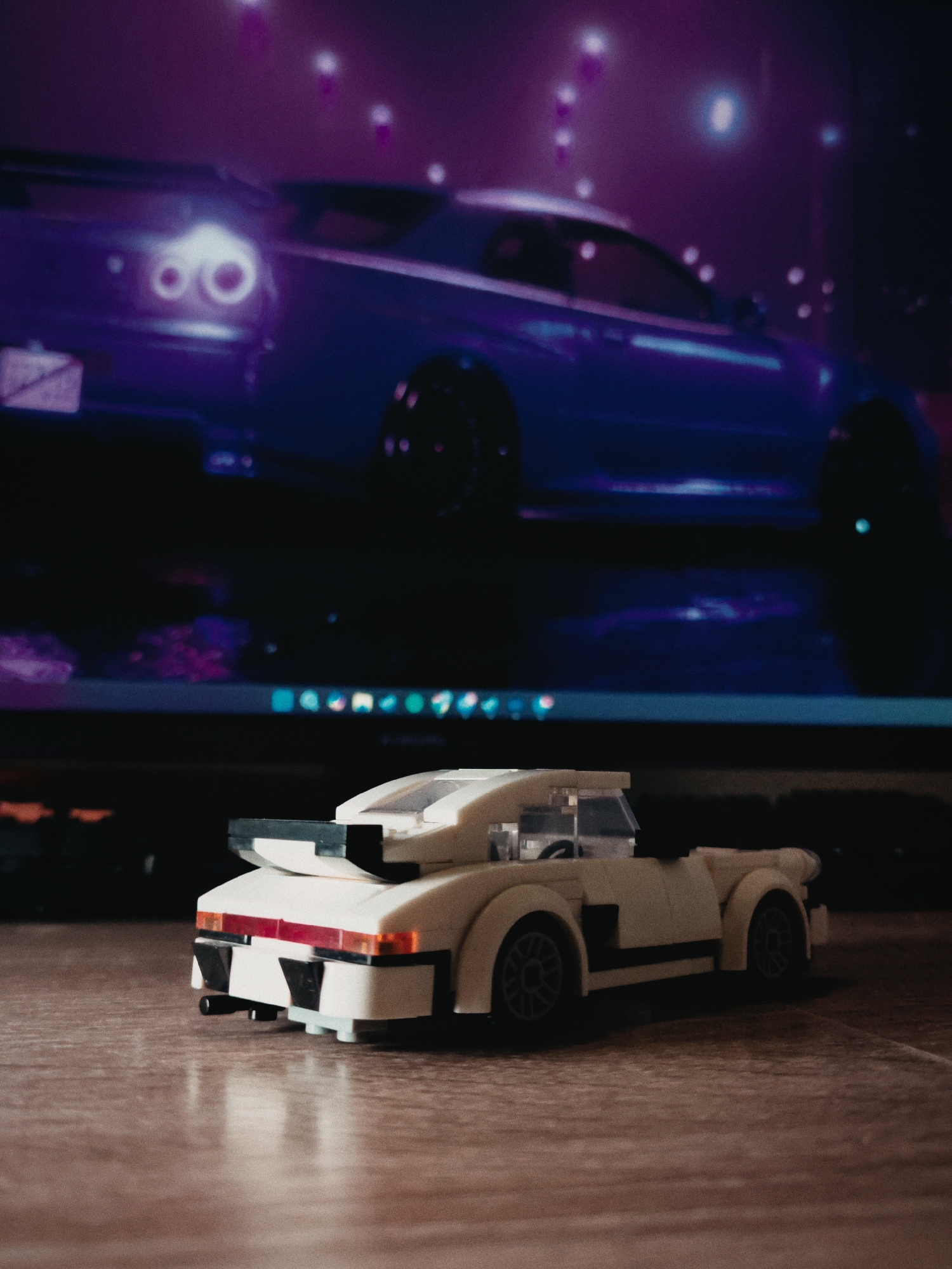 A LEGO car model sits on a wooden surface in front of a computer monitor displaying a blue sports car with distinctive circular taillights, likely a Nissan Skyline GT-R, set against a blurry background suggestive of a cityscape at night. The computer's taskbar is visible at the bottom of the screen. The background car's design features are consistent with those seen in promotional material relating to Budva, Montenegro; but this can't be confirmed to be the exact location of where the background image on the screen was taken.