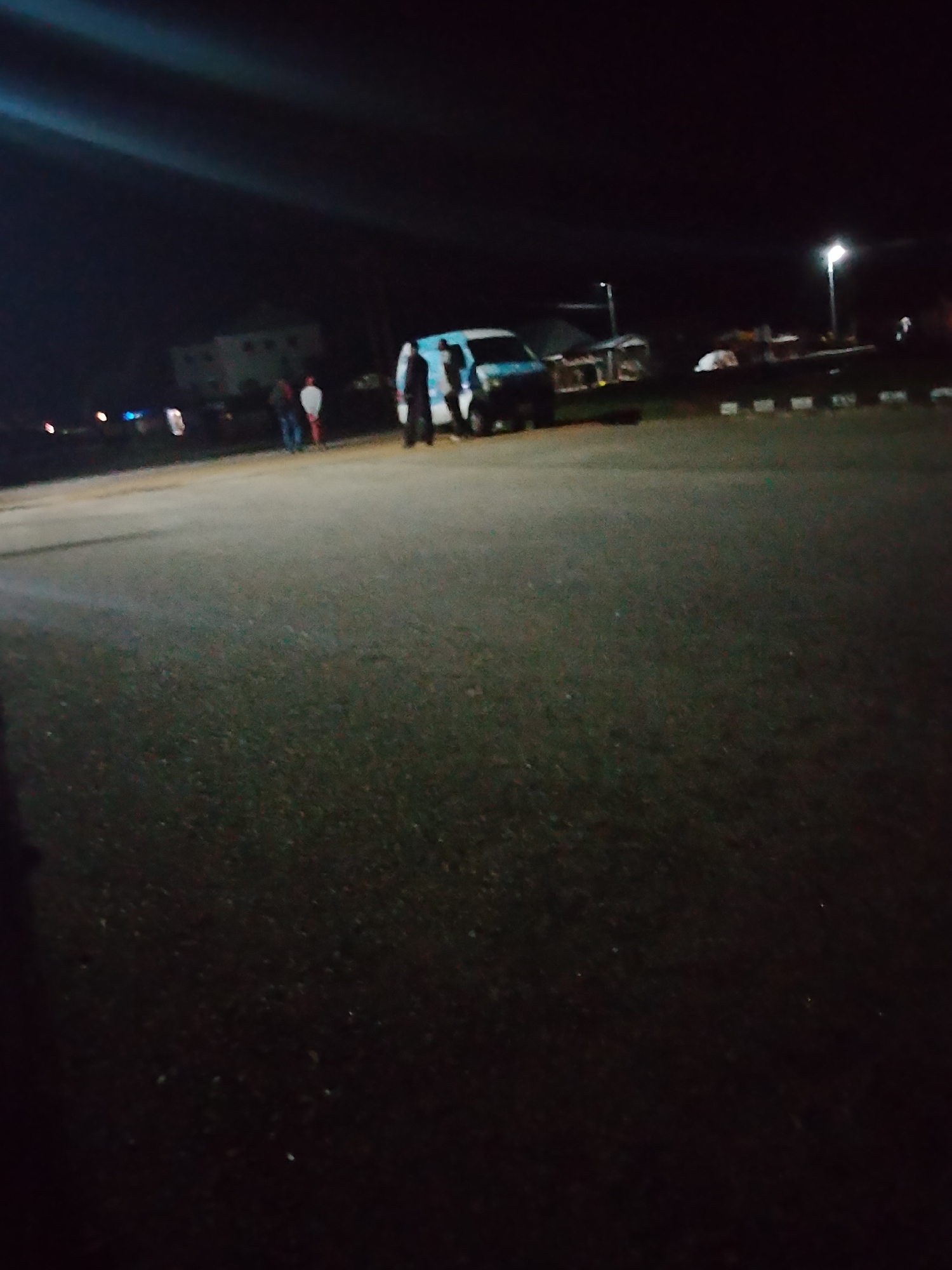 The image depicts an outdoor scene at night, likely in Jalingo, Nigeria. In the background, a multi-story building with illuminated windows is visible. Several individuals are standing near a white van with blue and white markings, possibly a police or utility vehicle. The van appears to be parked on an asphalt surface, which is the predominant feature of the foreground. The environment is dark, with artificial lighting from streetlights and the building illuminating parts of the scene. The overall impression is that of a public area, possibly a parking lot or street, at night.